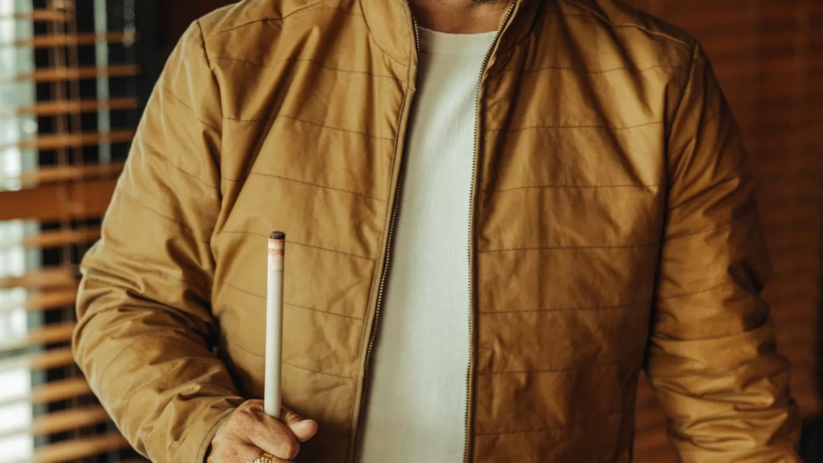 A model wearing a tan colored version of Flint and Tinder's York Quilted Waxed Jacket. The image is cropped to show the models torso between his wait and shoulders. The jacket is unbuttoned to reveal a white cream shirt. A pool cue is held in the models right hand.