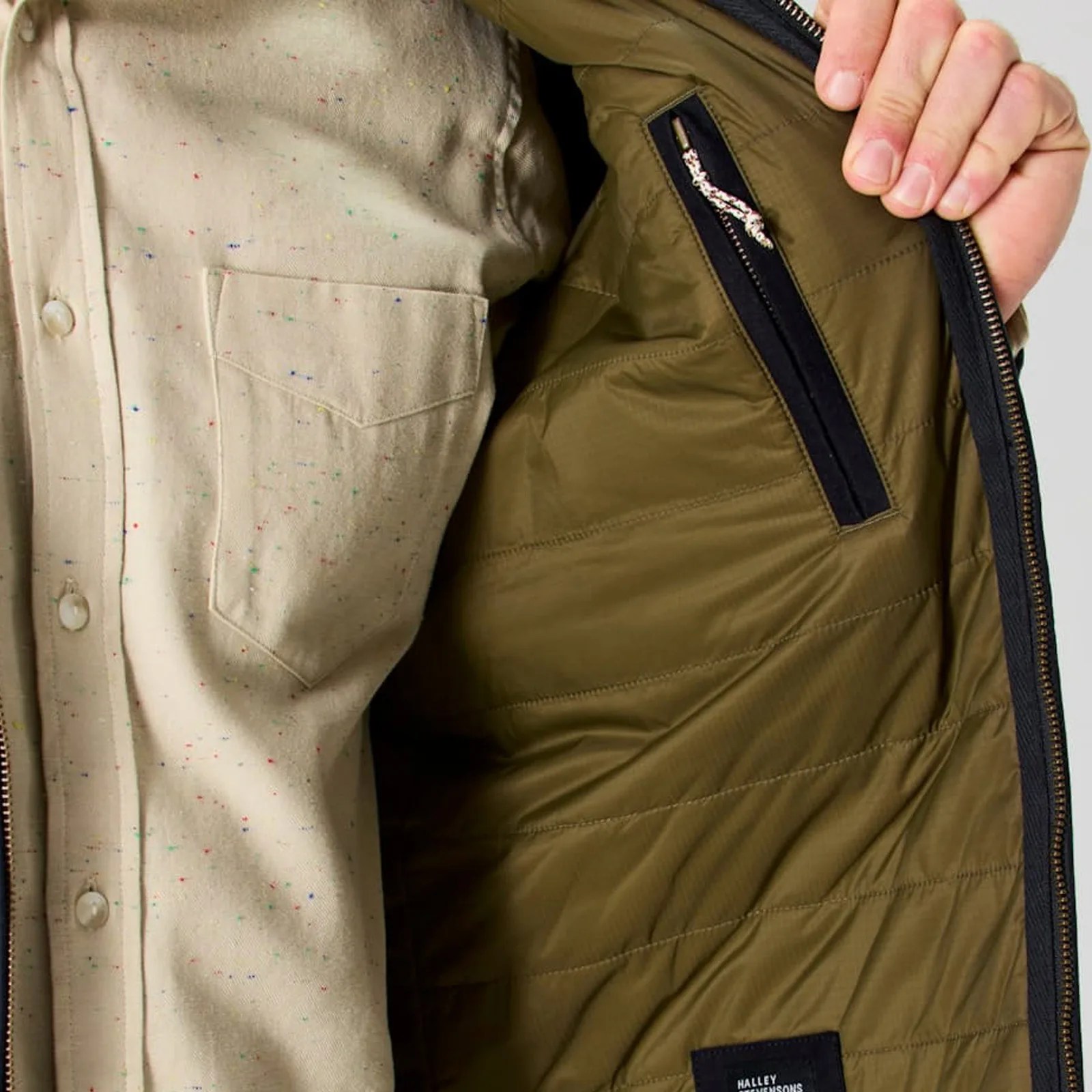 A photo showing the interior zippered chest pocket of the Flint and Tinder York Quilted Waxed Canvas Jacket. The interior lining of the jacket is a brownish green. The models shirt is a speckled light cream color. The models hand is pulling on the jacket's zippered edge to reveal the jacket's left interior and pocket. 