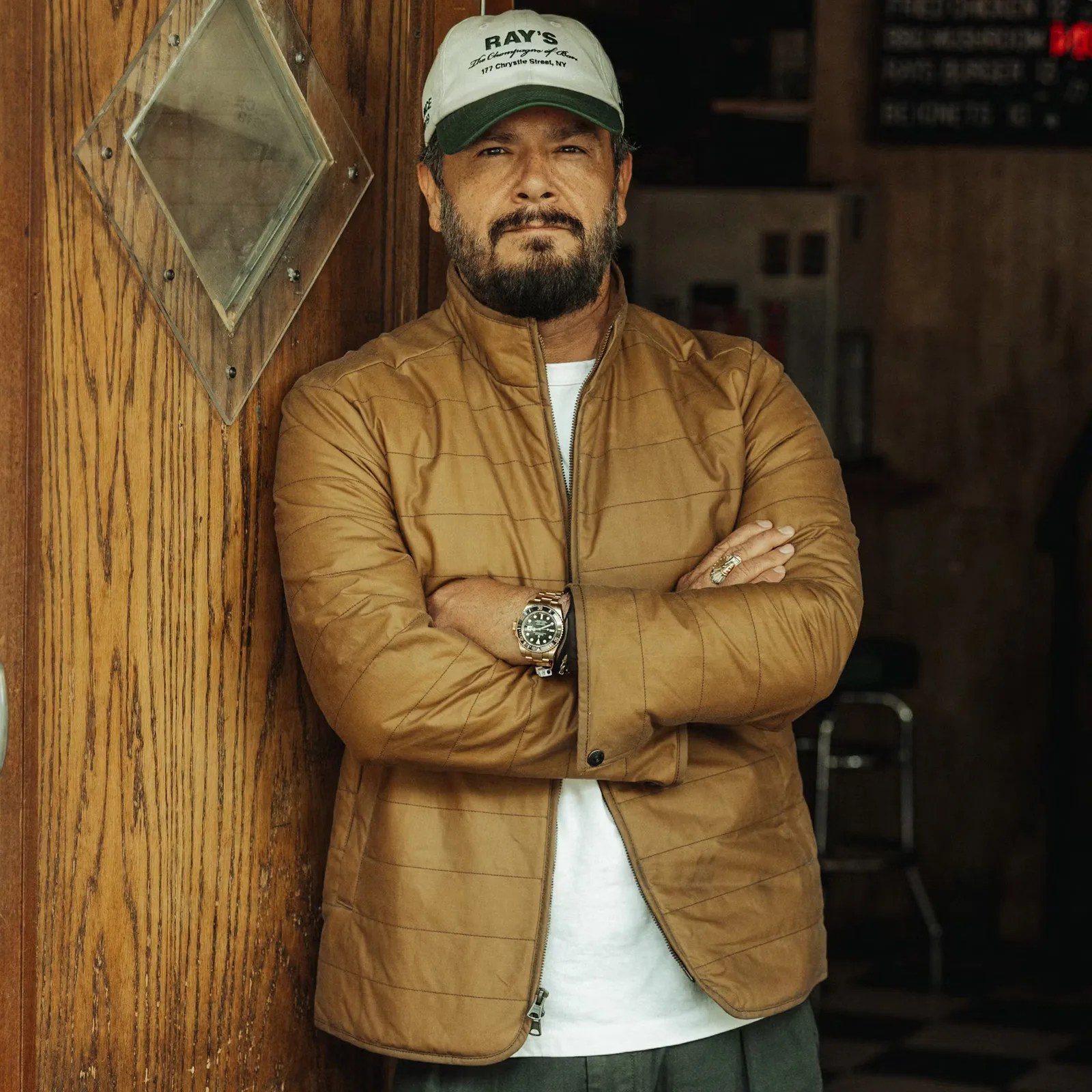 A bearded man directly facing the camera with his arms crossed wearing Flint and Tinder's York Quilted Waxed Jacket in tannish brown color. He is  wearing a baseball hat and white t-shirt underneath the jacket. He is leaning against a wooden bar door with a diamond shaped window cut into the upper portion. Blurred details of a bar including a stool and menu board appear in the background. 