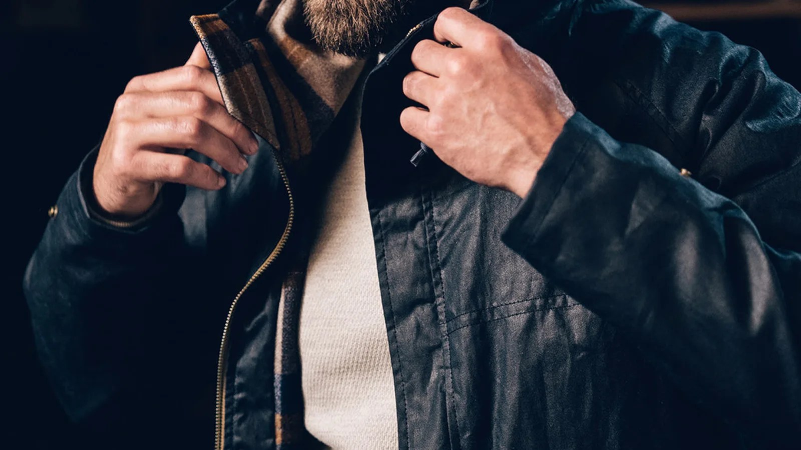 A close up of the chest area of a man wearing the Filson x Freeman Wool Lined Raincoat. The mans arm's are shown pulling up the jacket's hood. The jacket is a black waxed canvas. The shirt underneath the jacket is a light off-white.