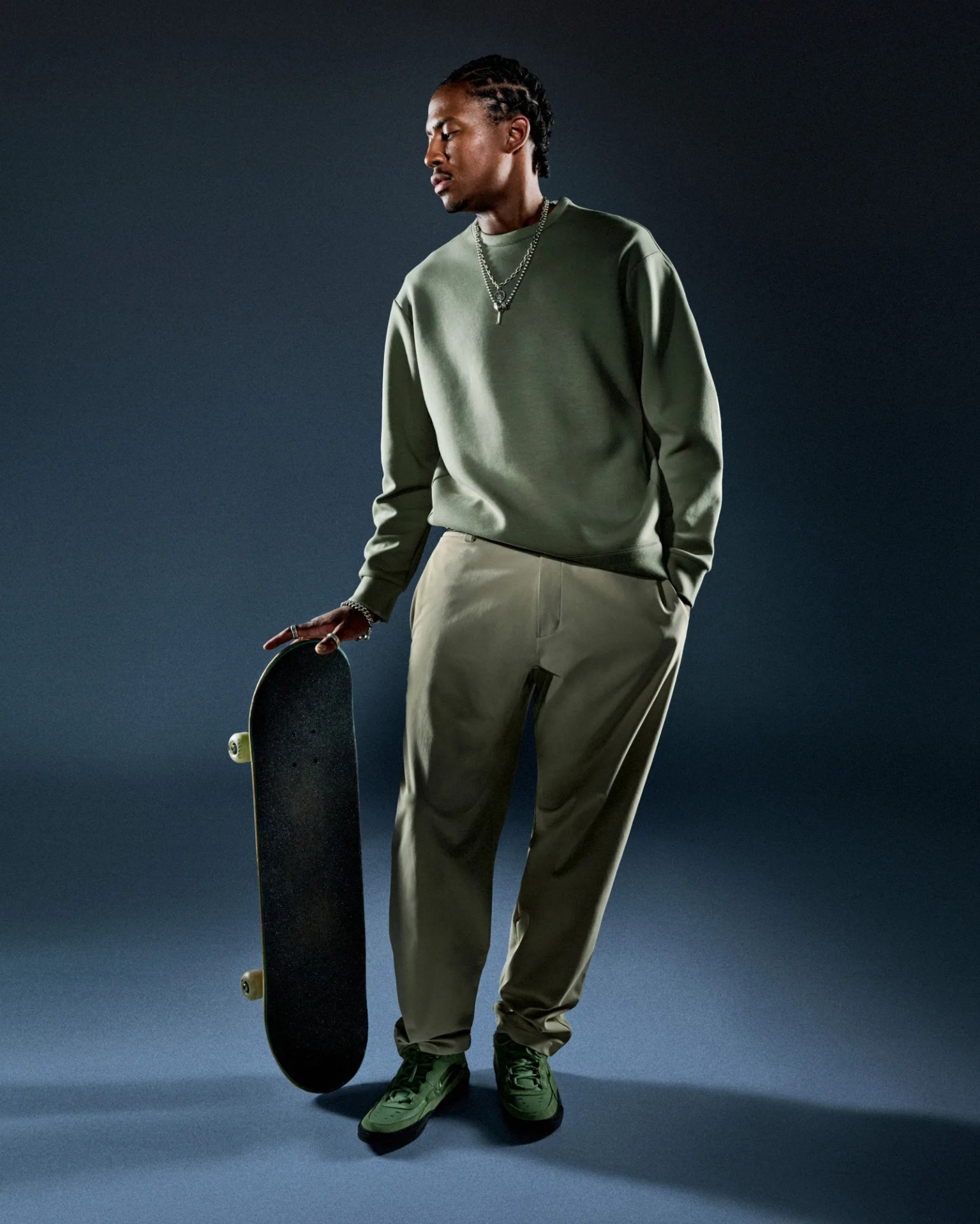 A black male model wearing a sweat shirt and khaki pants from Nike's 24.7 collection. He is shown standing and looking to the left. A skateboard is propped on the ground balanced under his hand. 