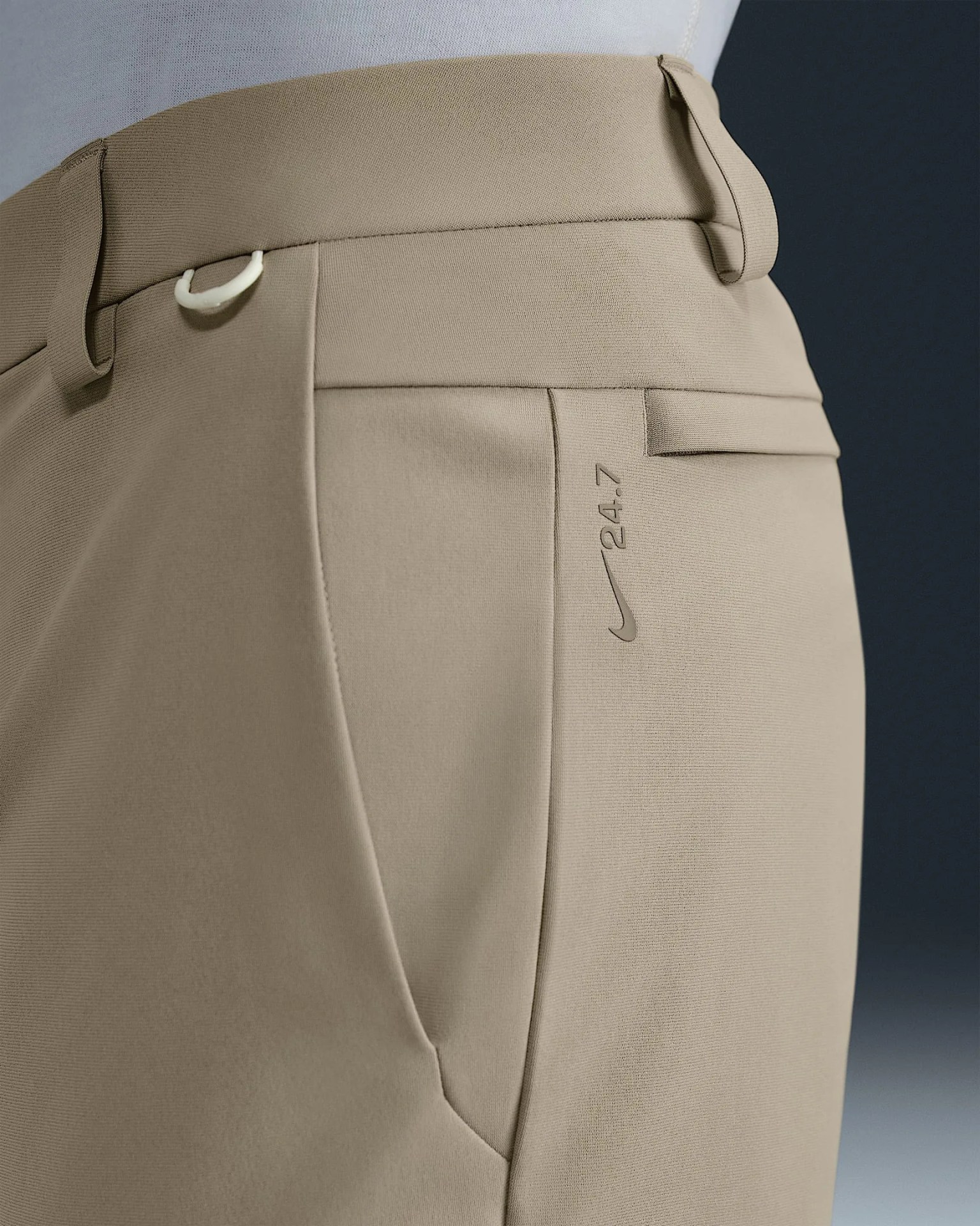 A close up of the belt and right pocket area of khaki pants included in Nike’s 24.7 collection. The pants are light tanned color. A darker Nike Swoosh with a 24.7 logo are visible right behind on the back edge of the pocket. 