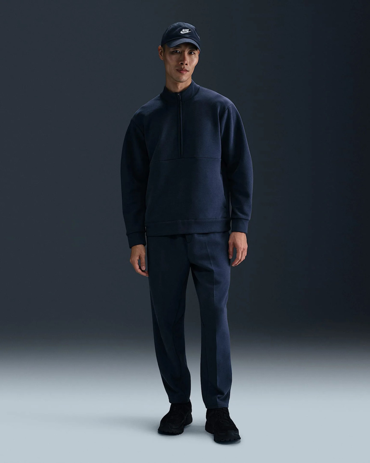 A male model wearing a navy colored quarter zip sweat shirt and pants from Nike’s 24.7 clothing collection. He is facing forward in the image and shown from head to toe. The background consists of an empty room with a black wall and light floor. 