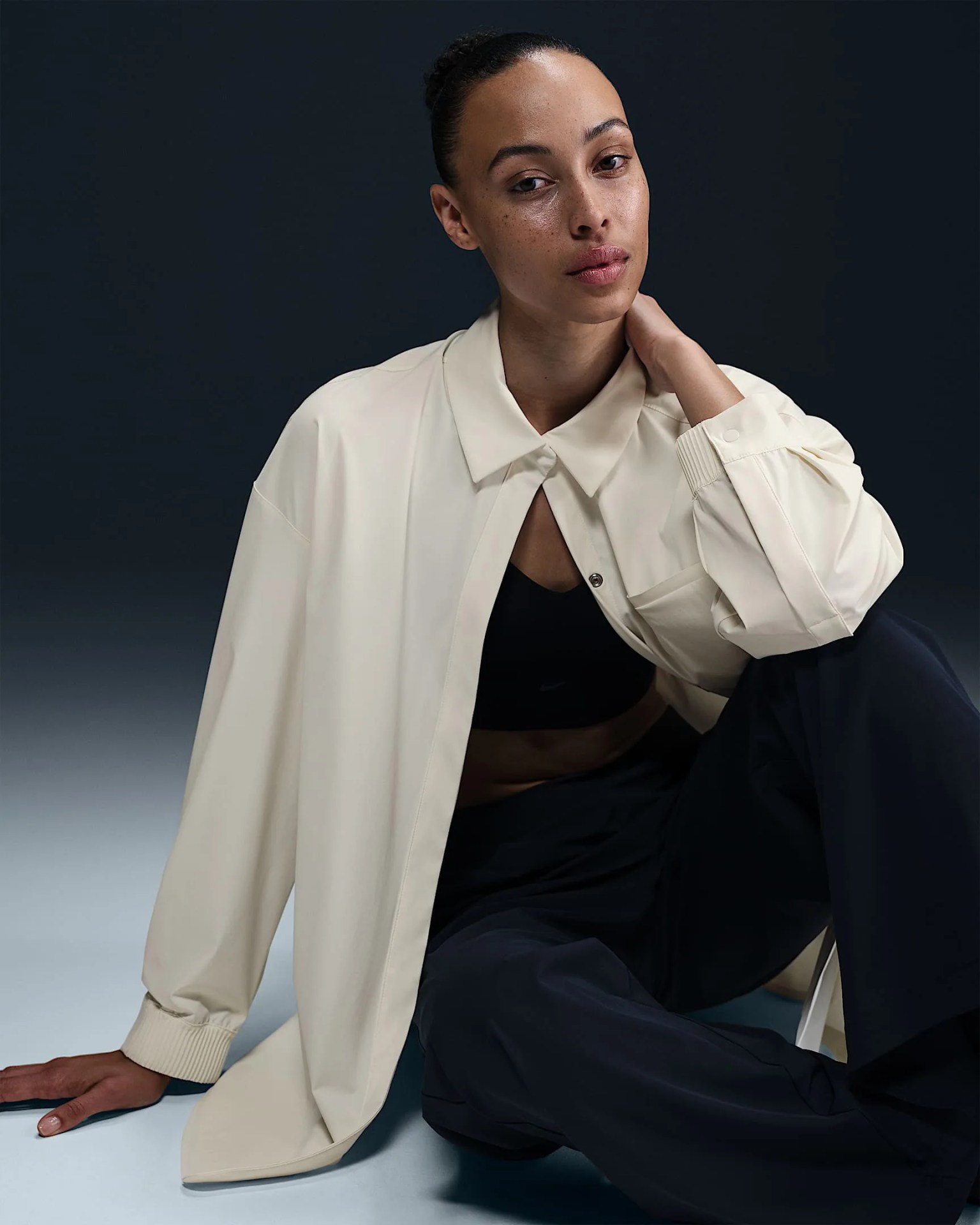 A female model wearing a cream white button up shirt from Nike’s 24.7 clothing collection. She is sitting down with her legs positioned in front. her right elbow is sitting against her right knee, propping her hand against her neck. The cream shirt is opened and unbuttoned except for the top button. The model is wearing a black sports bra and dark pants underneath. 