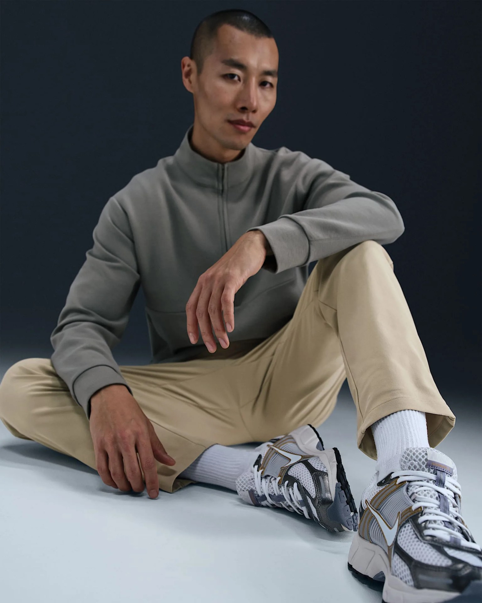A male Asian model shown wearing a grey quarter zip sweat shirt and tan chino pants that are part of Nike’s 24.7 collection. The man is shown seated on the ground with his legs in front. One leg is pulled closer to his body, while the other is extended and resting an angle. The man’s left arm and elbow are resting on his knee. He is staring directly ahead into the camera. The background is dark grey. The floor is light white. 