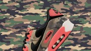 A Nike Air Max 90 is camo and red against a camo patterned background.