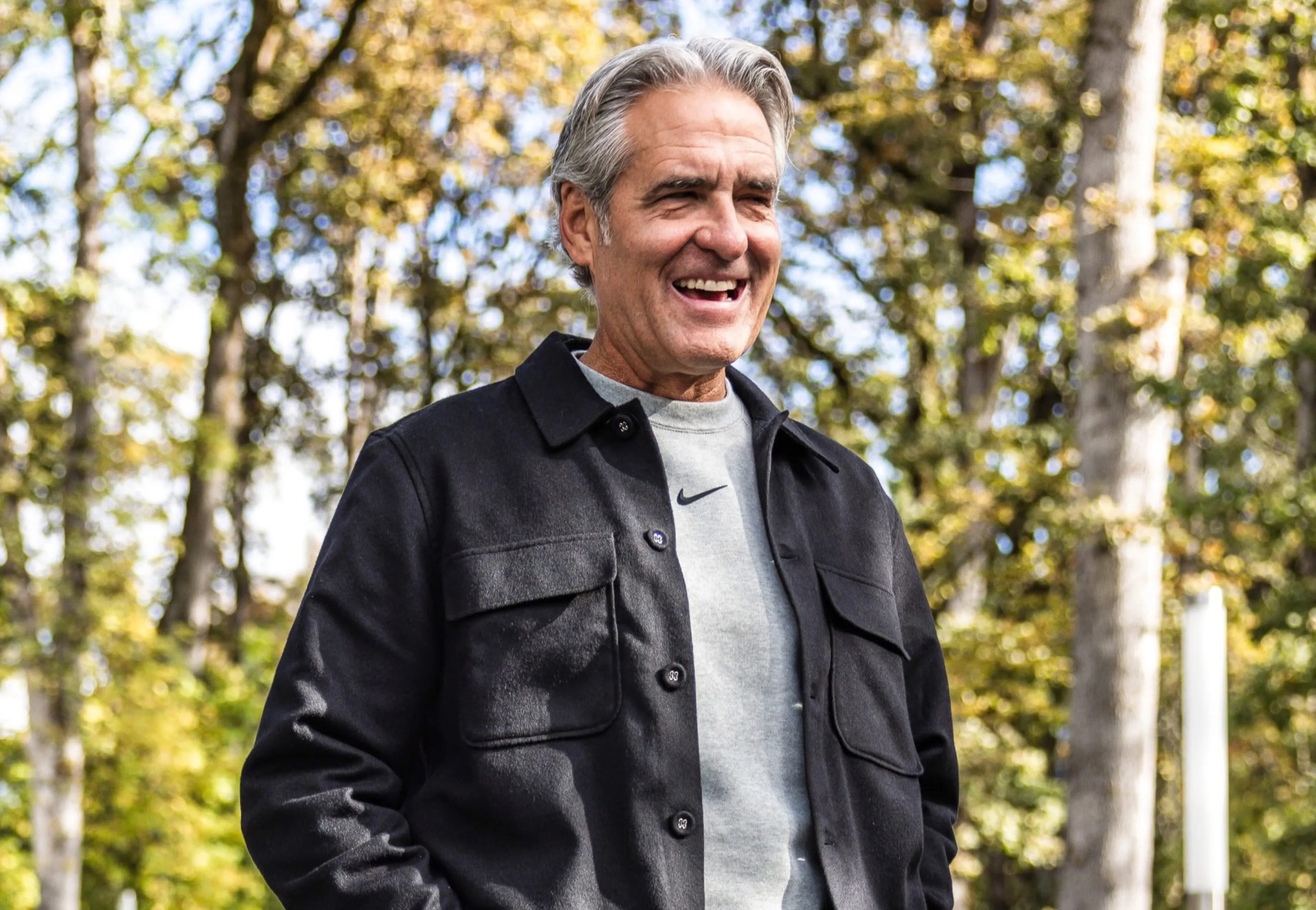 This is a portrait of Nike CEO Elliot Hill smiling with his mouth open, looking to the right. Hill is wearing a dark grey overshirt over a grey sweatshirt with a Nike Swoosh. Blurred trees appear around him in the background. 
