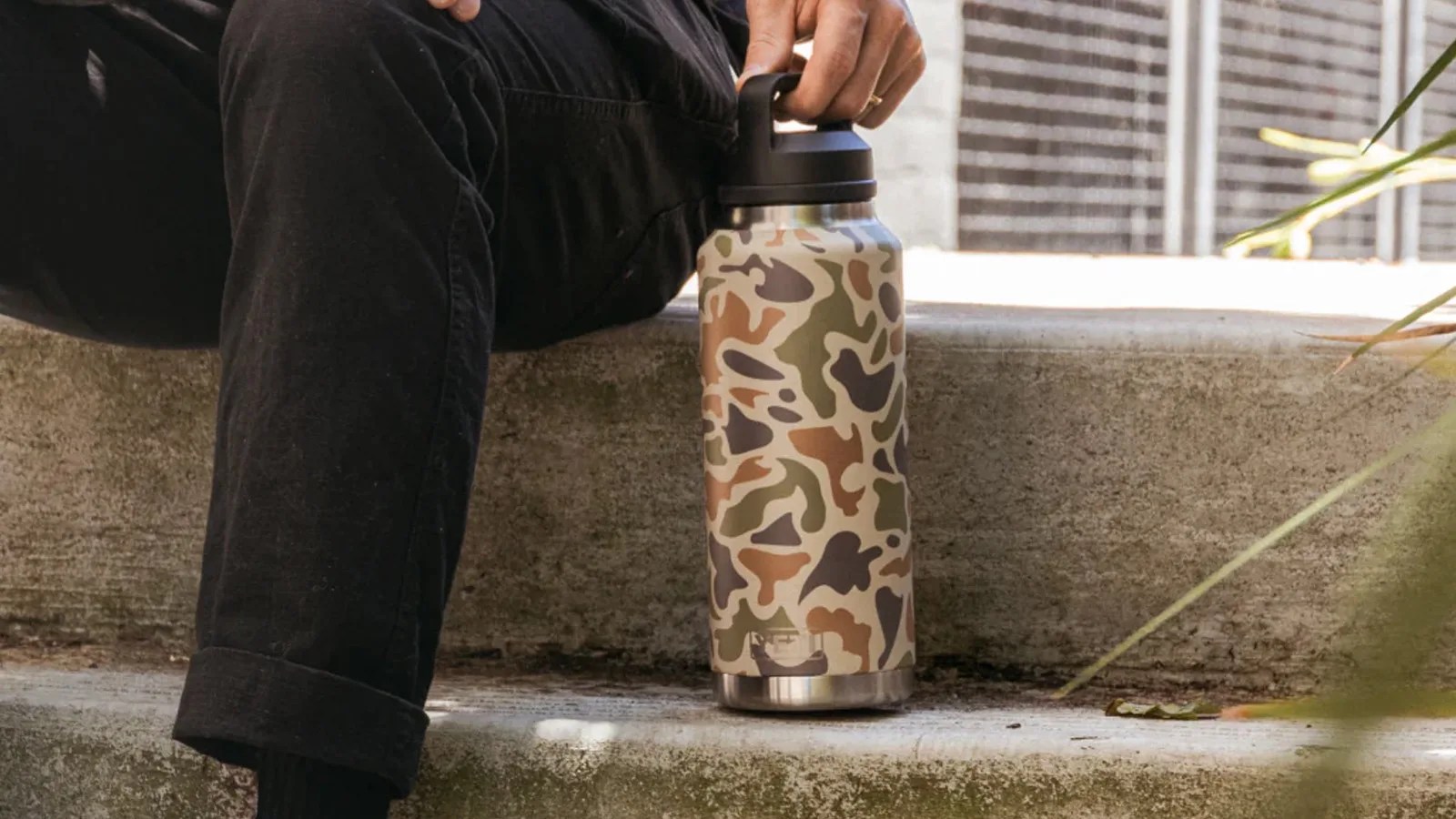 A Yeti Rambler in Tan Camo sitting on a concrete step next to someone sitting on a concrete step.