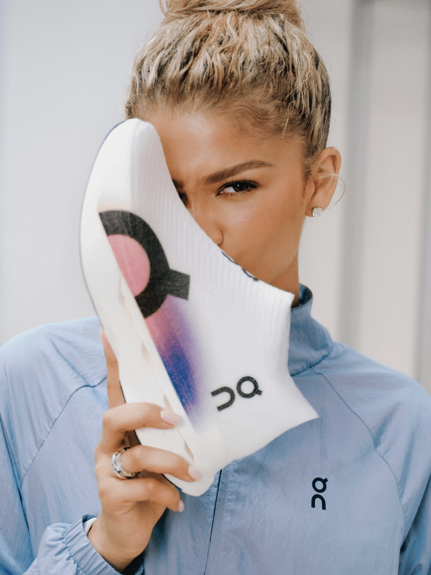 Zendaya shown holding up On Runnings LightSpray sneaker in one hand. The toe box of the shoe she is holding is covering one side of Zendaya's face, obscuring her right eye and right cheek. Zendaya is wearing a light blue track suit. 