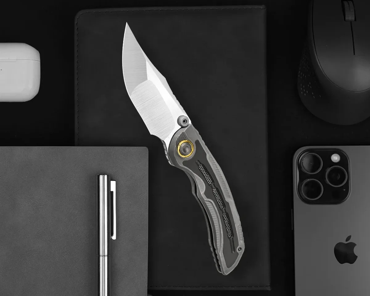 Kizer Silverwing knife over black desk with mouse, airpods, pen, notebook, and iphone