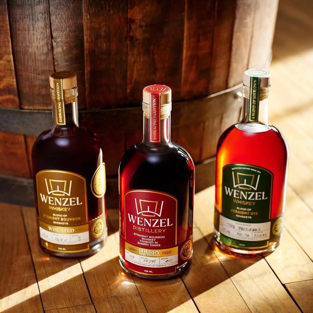 Three different bottles of Wenzel Whisky shown sitting on a distilery floor in front of a whiskey barrel. One bottle label is maroon, another is dark green, and another is gold. All bottles are backlit with sunlight, casting shadows in front of them. 