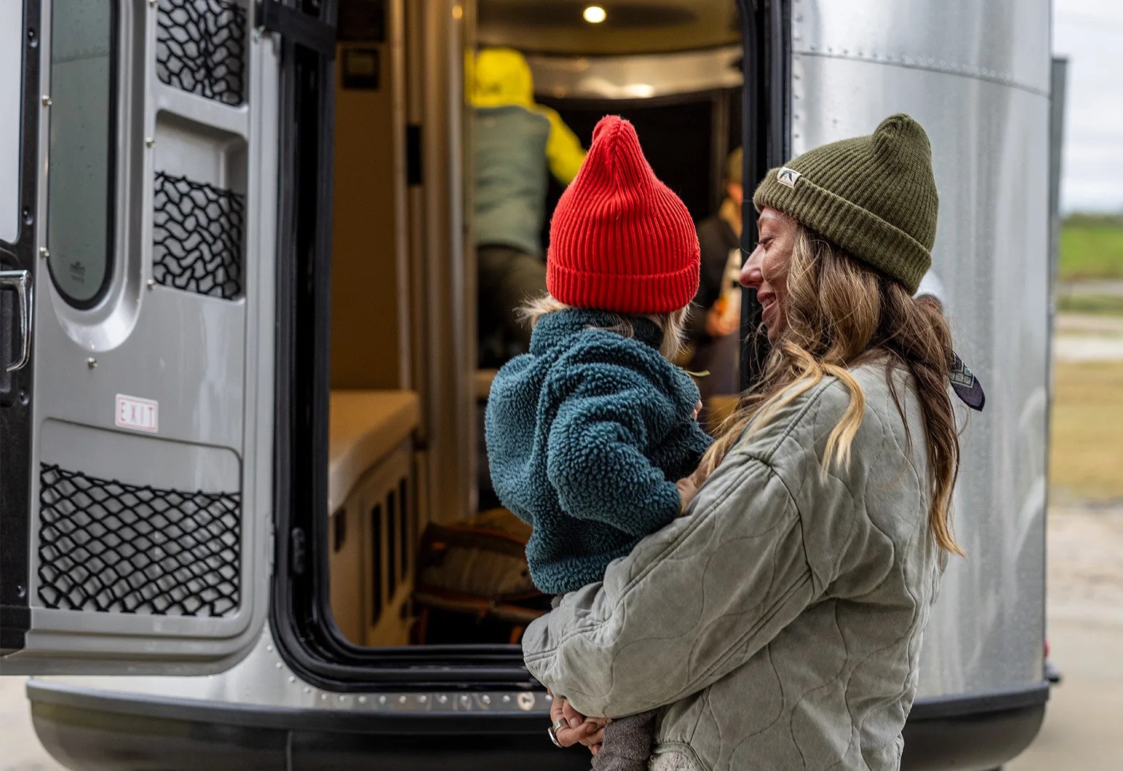 A woman holding a child. Both are dressed in fleeces and warm clothes. They are standing in front of the open rear door of the Airstream Basecamp Xe trailer