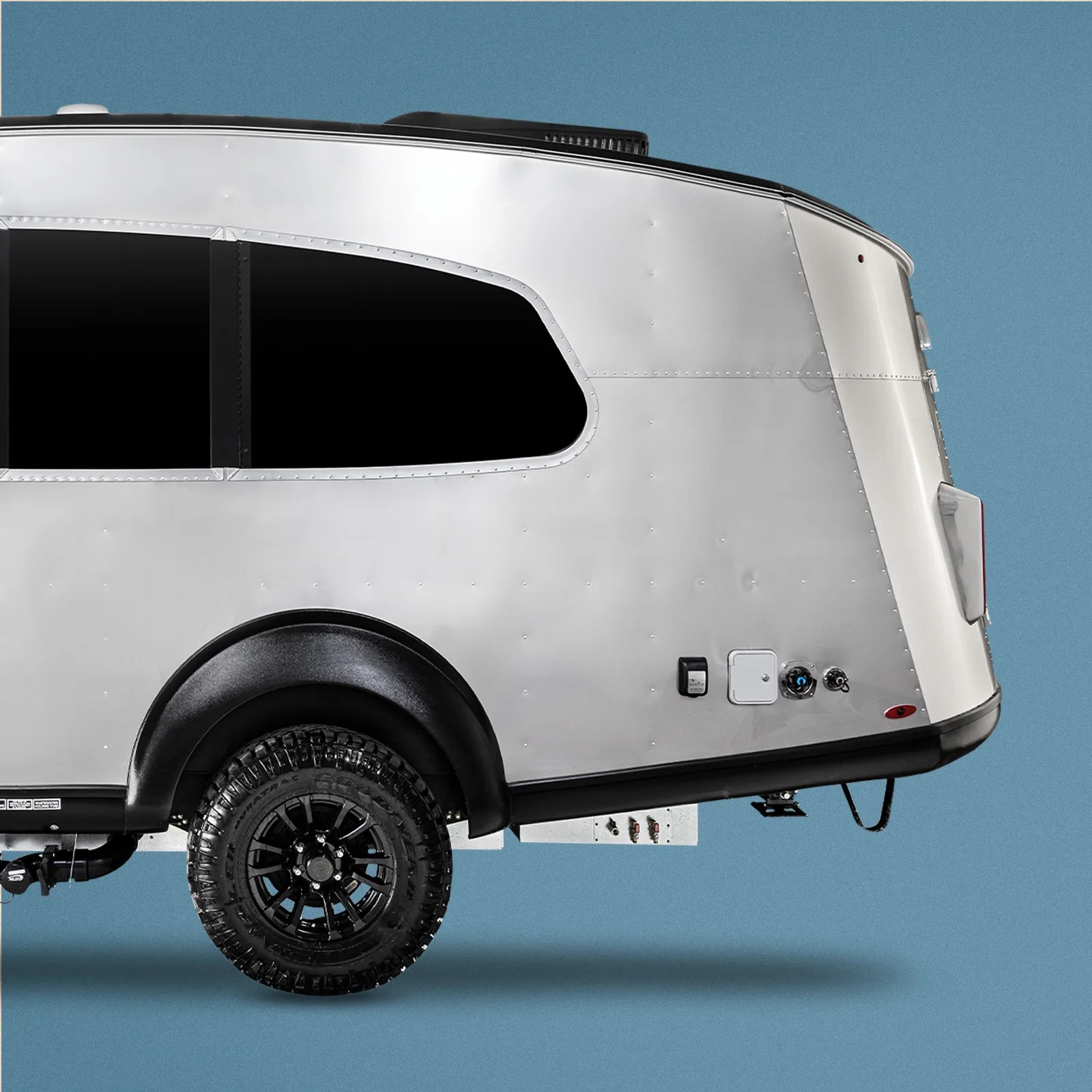 A close-up of the rear and wheel area of the Airstream Basecamp Xe adventure trailer. The trailer is shown against a grainy light blue background.