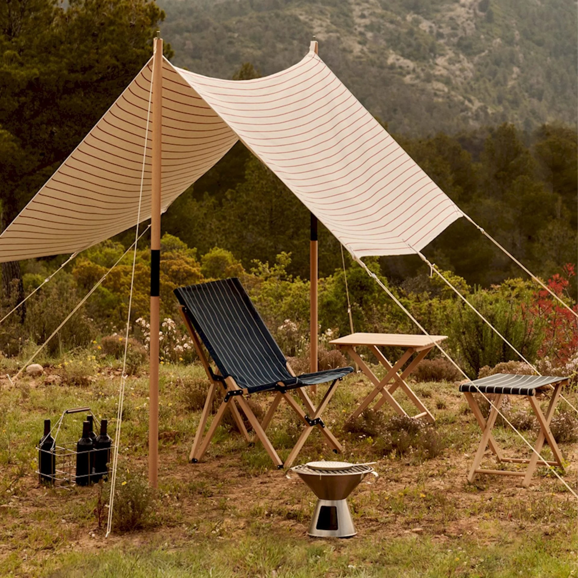 Beige striped canvas canopy with wooden poles over a black striped folding chair, wooden table, stool, and portable grill on grass.