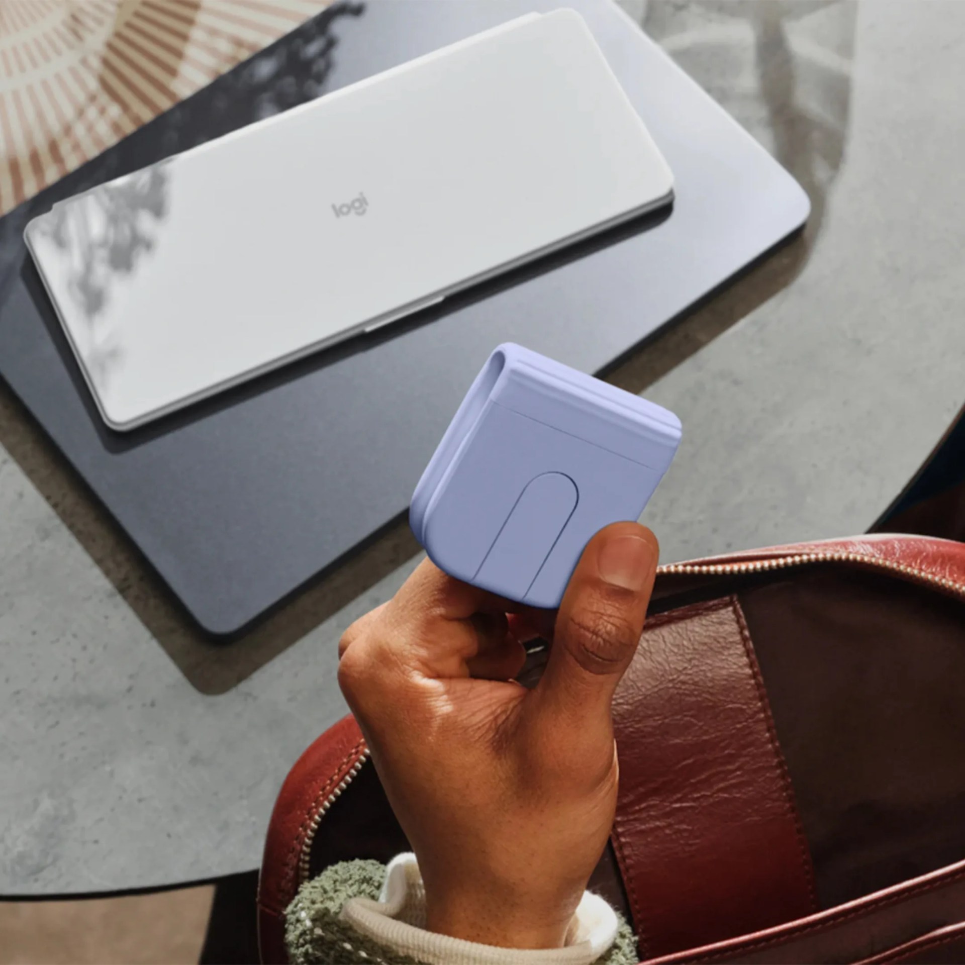 Hand holding a small, light purple device with a button, above a brown leather bag and two closed laptops.