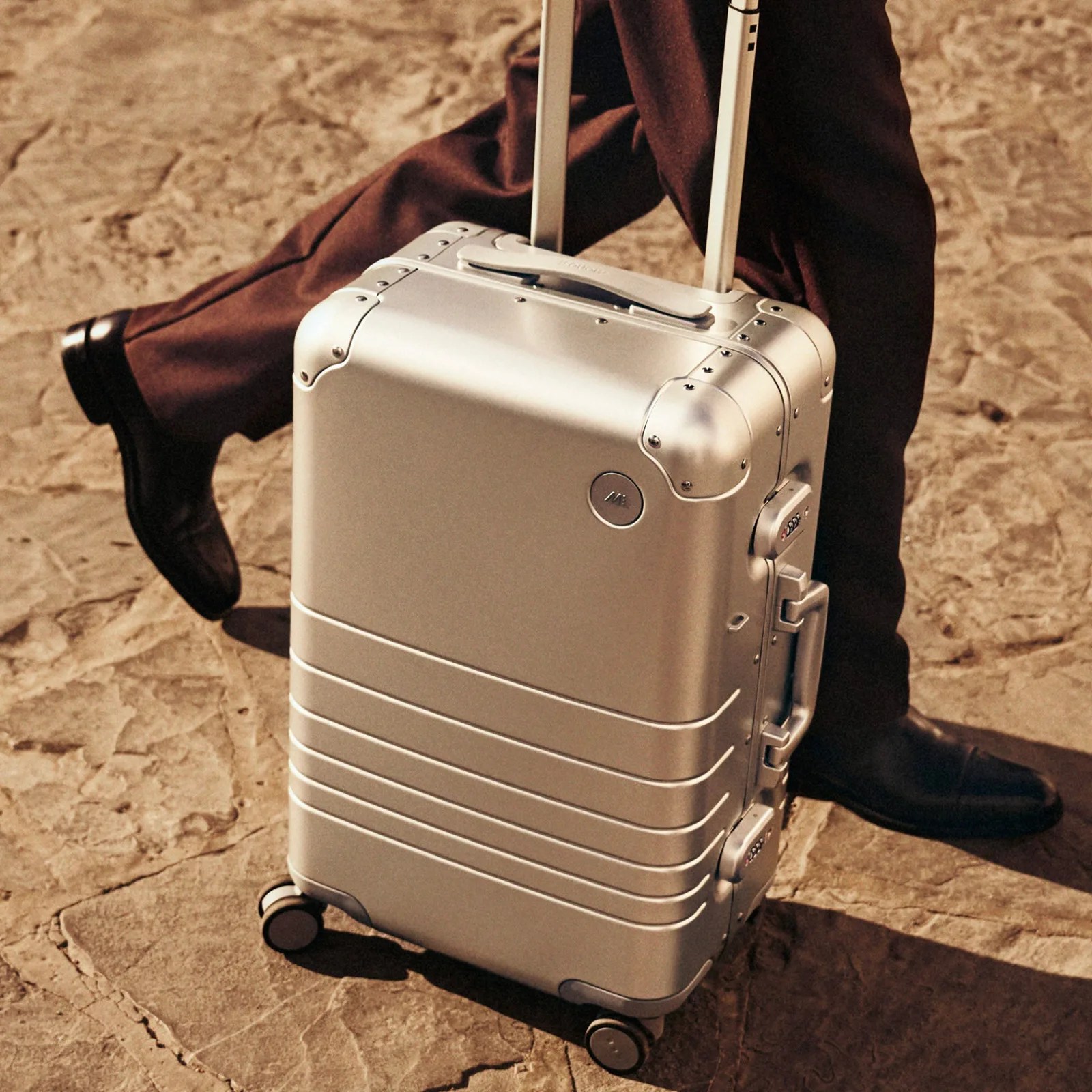 A Monos aluminum suitcase is being rolled on a stone street by a person wearing brand slacks and black shoes.