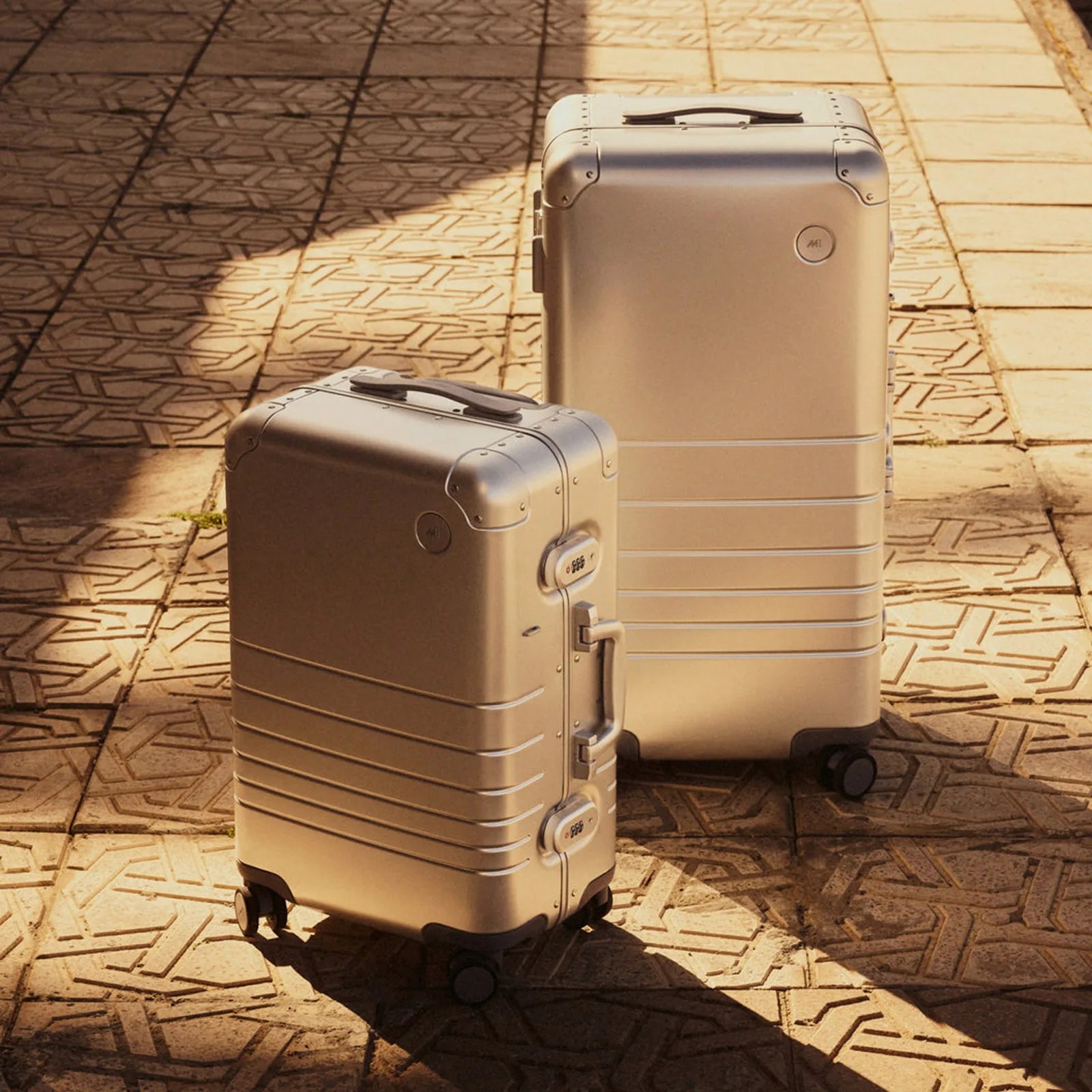 A small Monos aluminum suitcase standing at an angle and in front of a Monos aluminum trunk. Both bags are shown on a stone tile surface with interesting patterns on the floor. Warm sunlight and shadows cover both bags.