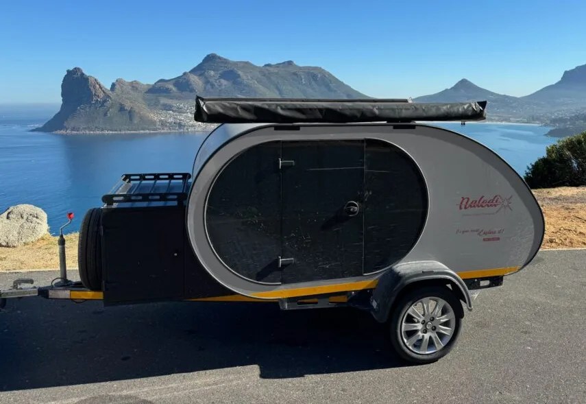 A Naledi Camper trailer shown parked in front of a large body of water with hills and mountains in the background. 