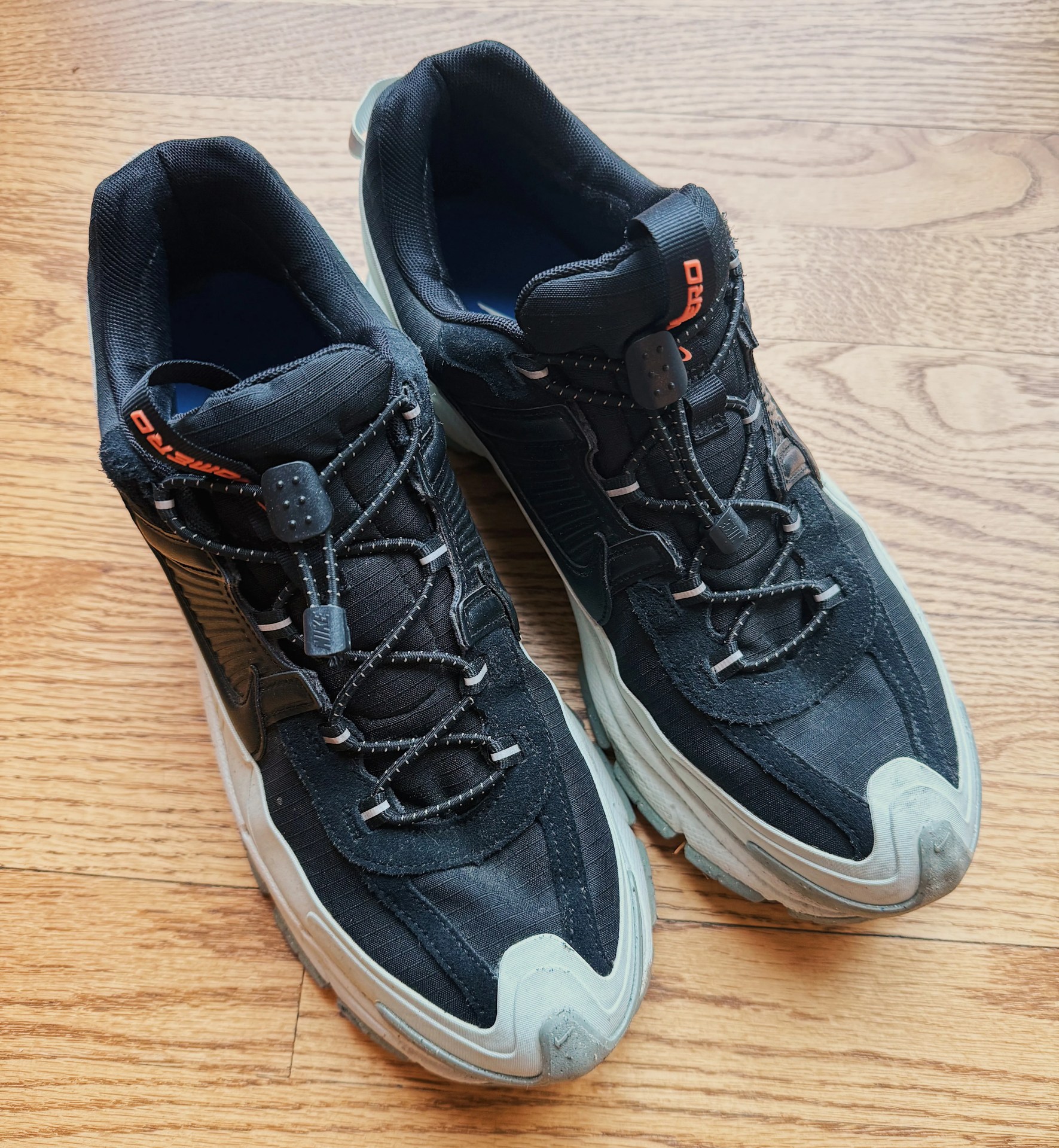 Pair of black and gray Nike sneakers with reflective laces and orange logo on wooden floor.