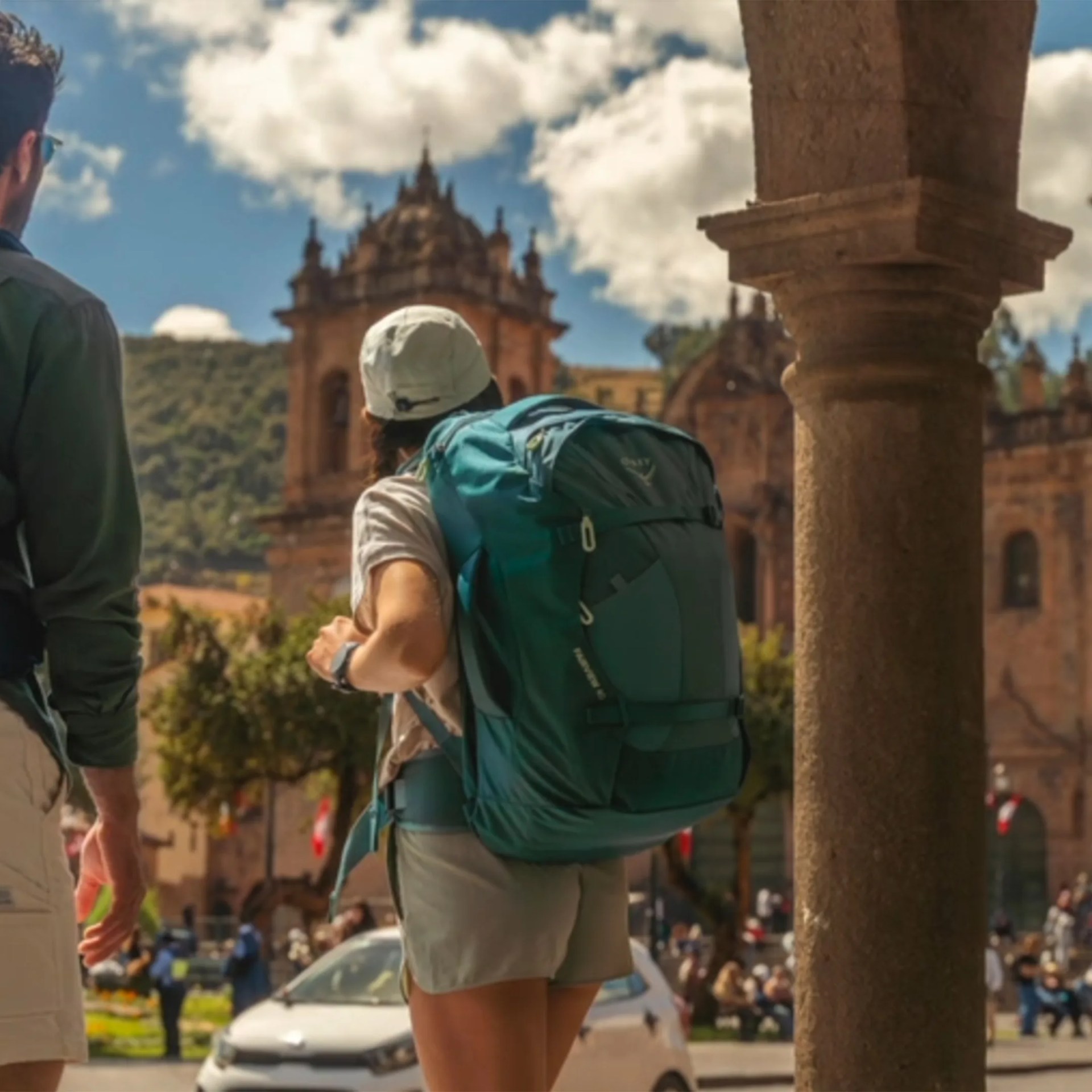Person wearing a large teal hiking backpack and white cap standing near a stone column with historic buildings in the background.