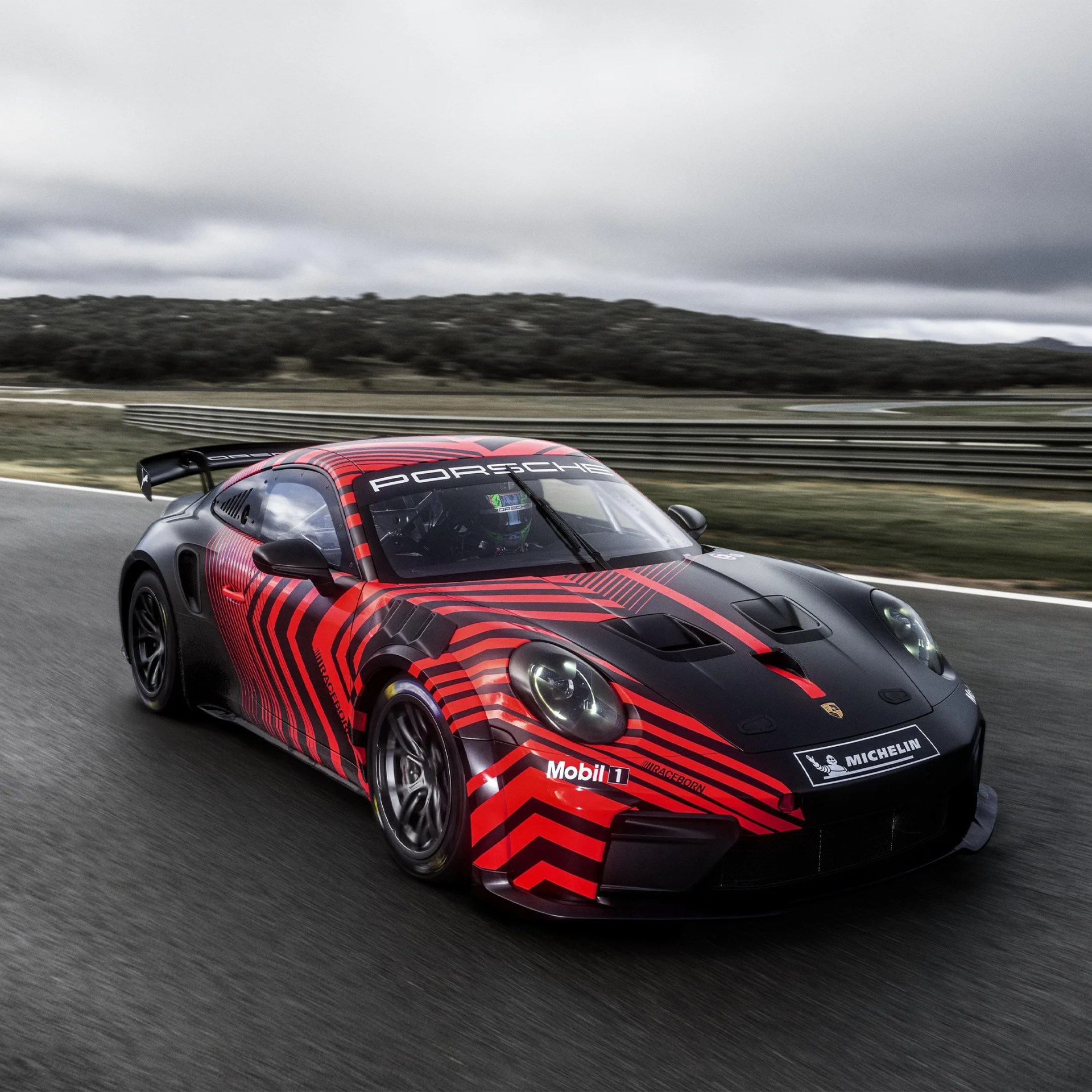 Black and red Porsche race car with racing decals on a track under cloudy sky.