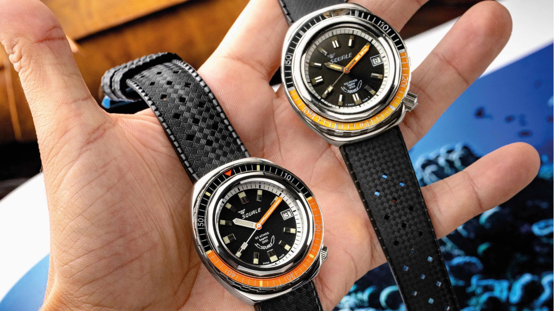 An original and new version of the Squale 2001 dive watch held in one hand with an open palm. The vintage version sits on the right hand side towards the fingers, while the new version is placed in the palm of the hand. Both watches feature bezels that are half black, half bright orange and black watch faces. Both also feature rubber tropical style straps. 