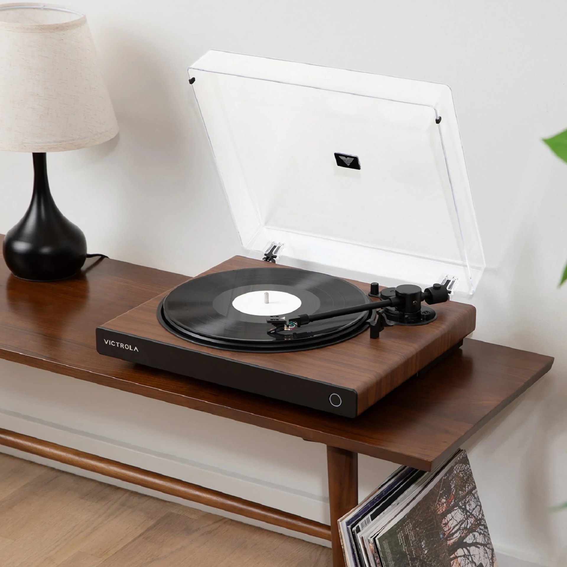 Wooden Victrola turntable with clear dust cover and black vinyl record on a wooden table next to a black lamp with a beige shade.