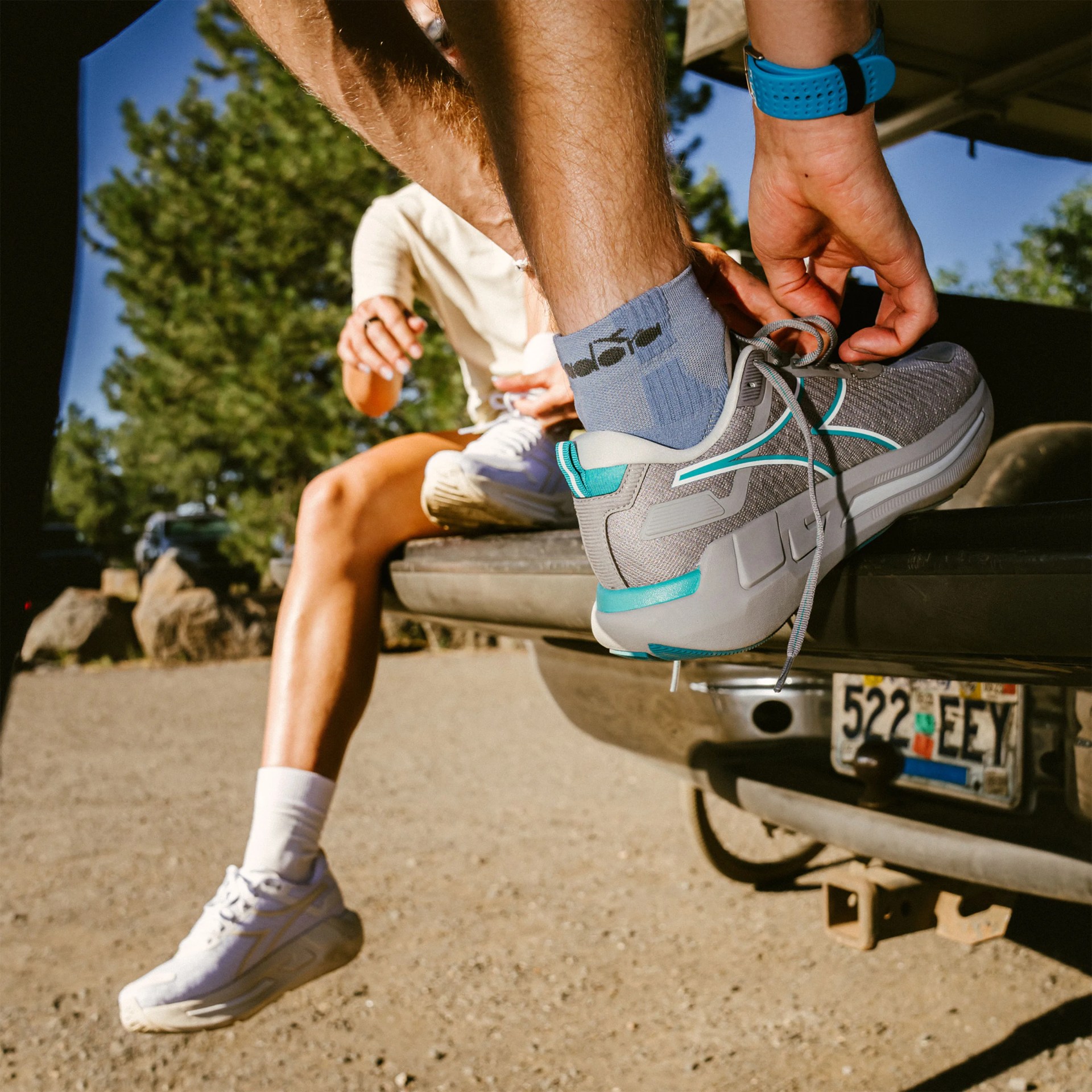 Person tying gray and teal running shoes while sitting on the tailgate of a vehicle with a visible license plate.