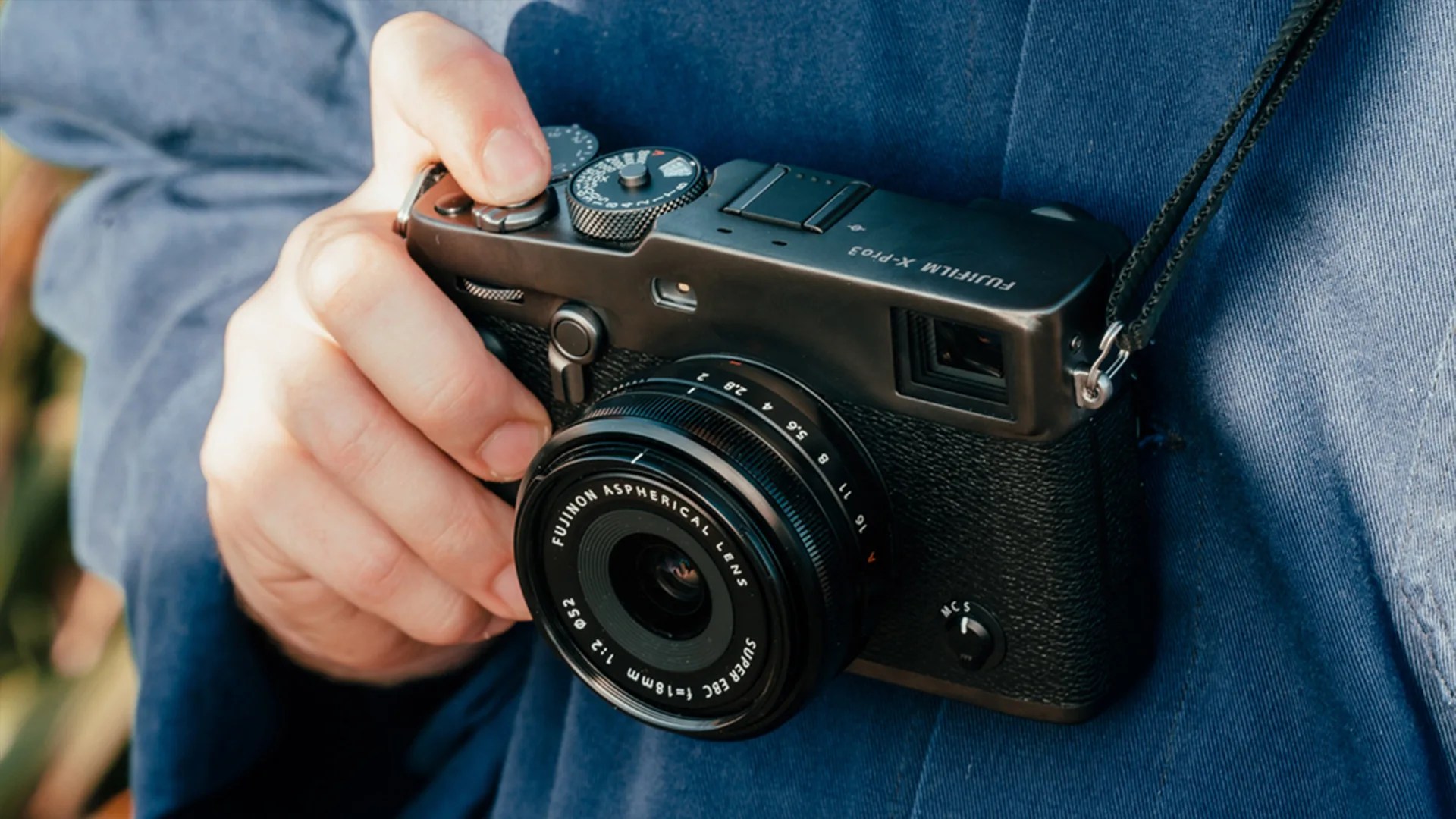 Black Fujifilm X-Pro3 camera with Fujinon 18mm f/2.0 lens held in a hand against a blue fabric background.