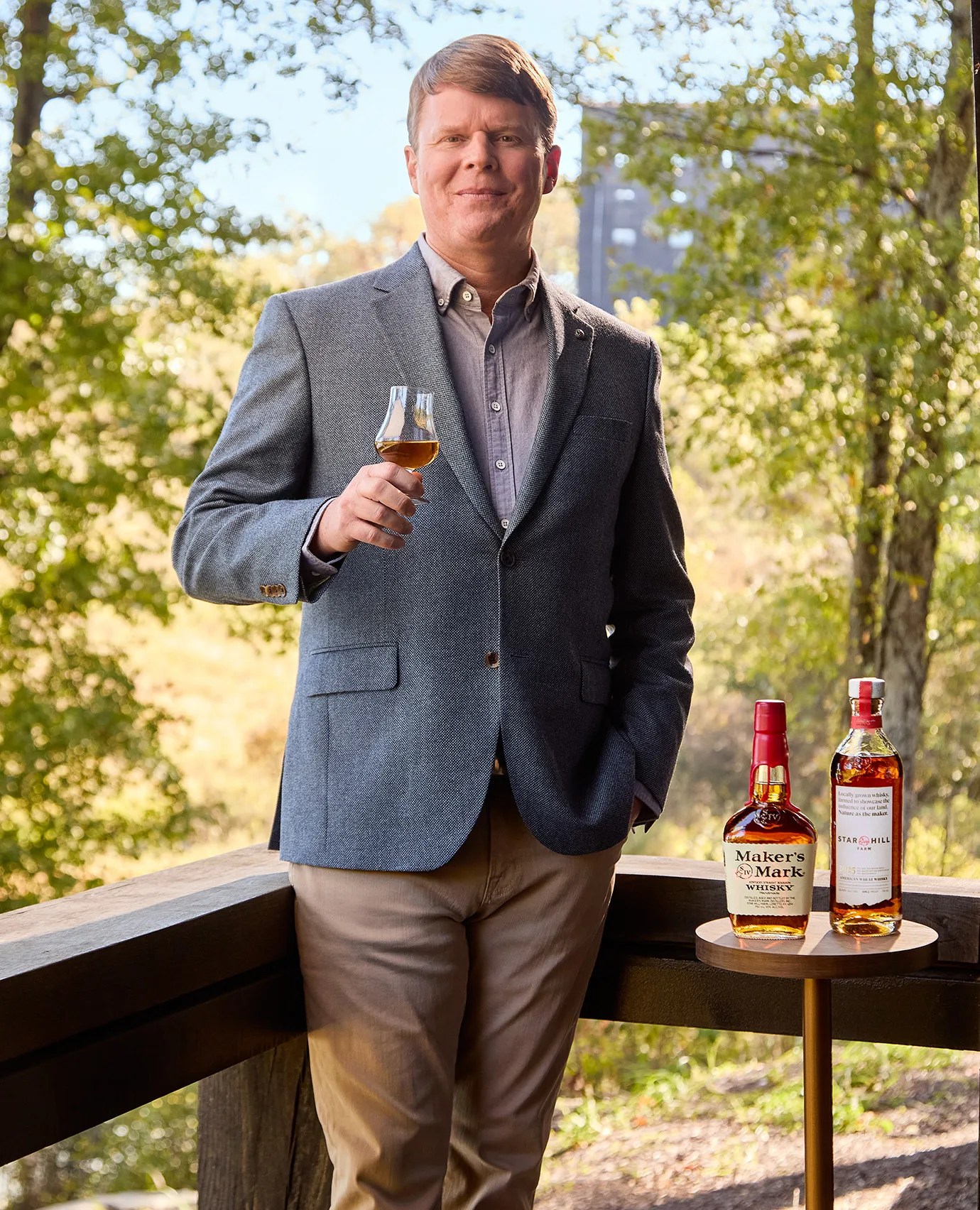 A portrait photo of Rob Samuels, the 8th generation distiller and managing director of Maker's Mark Distillery shown holding a glass of whisky standing next to a bottle of Maker's Mark and Star Hill Farms Whisky