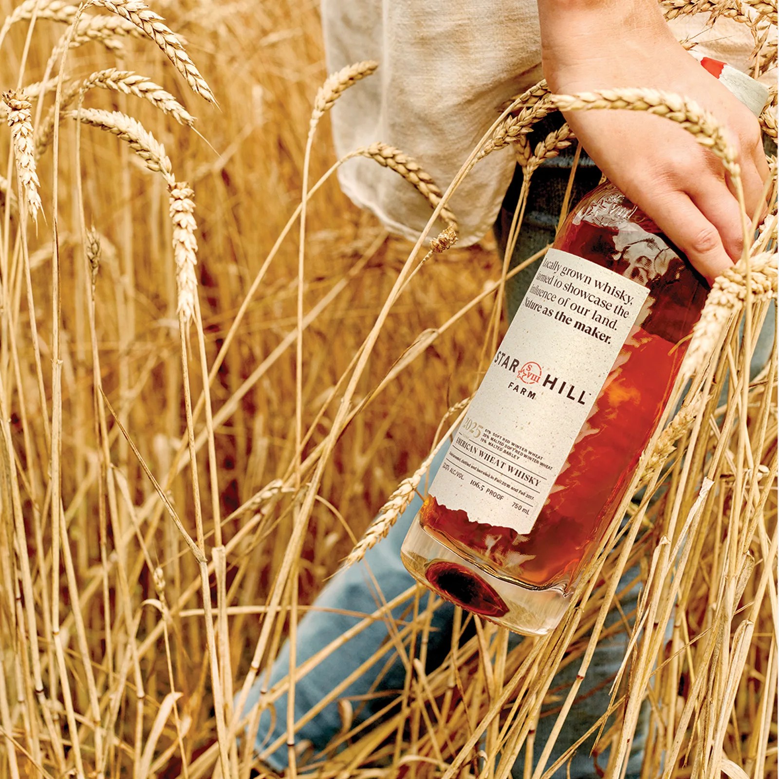 A bottle of Star Hill Farm Whisky shown being carried in one hand by a person walking through a field of golden wheat.