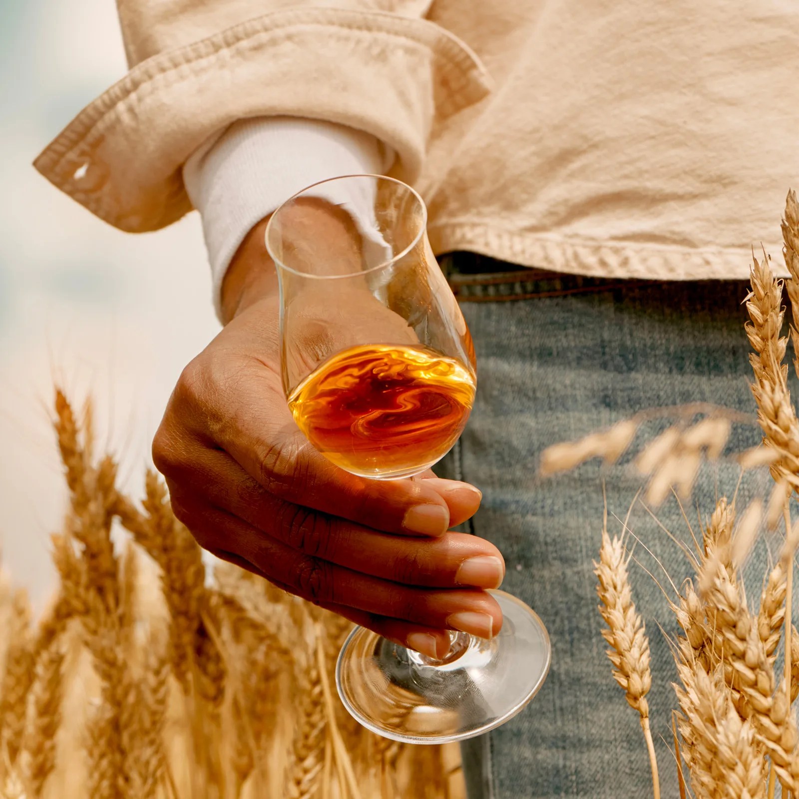 A glass of Star Hill Farm Whisky held in the hand of a model standing in a wheat field shown from the waist down to the thigh.