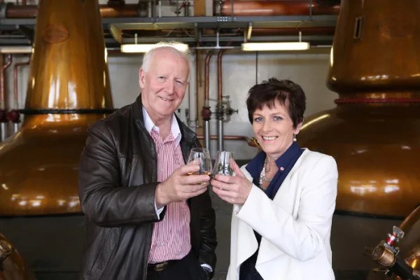 The GlenAllachie owners Billy Walker and Trisha Savage shown holding up glencarin of whisky in front of two copper whisky stills.