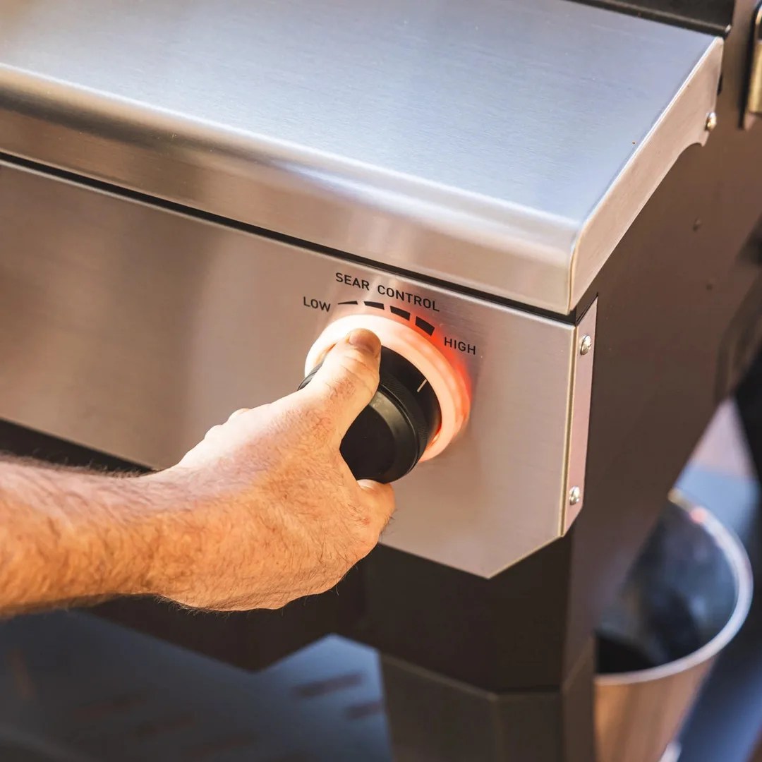 A hand is shown adjusting the sear control knob on the Recteq X-Fire Pro 825 pellet grill. The knob is shown illuminated with an orange ring.