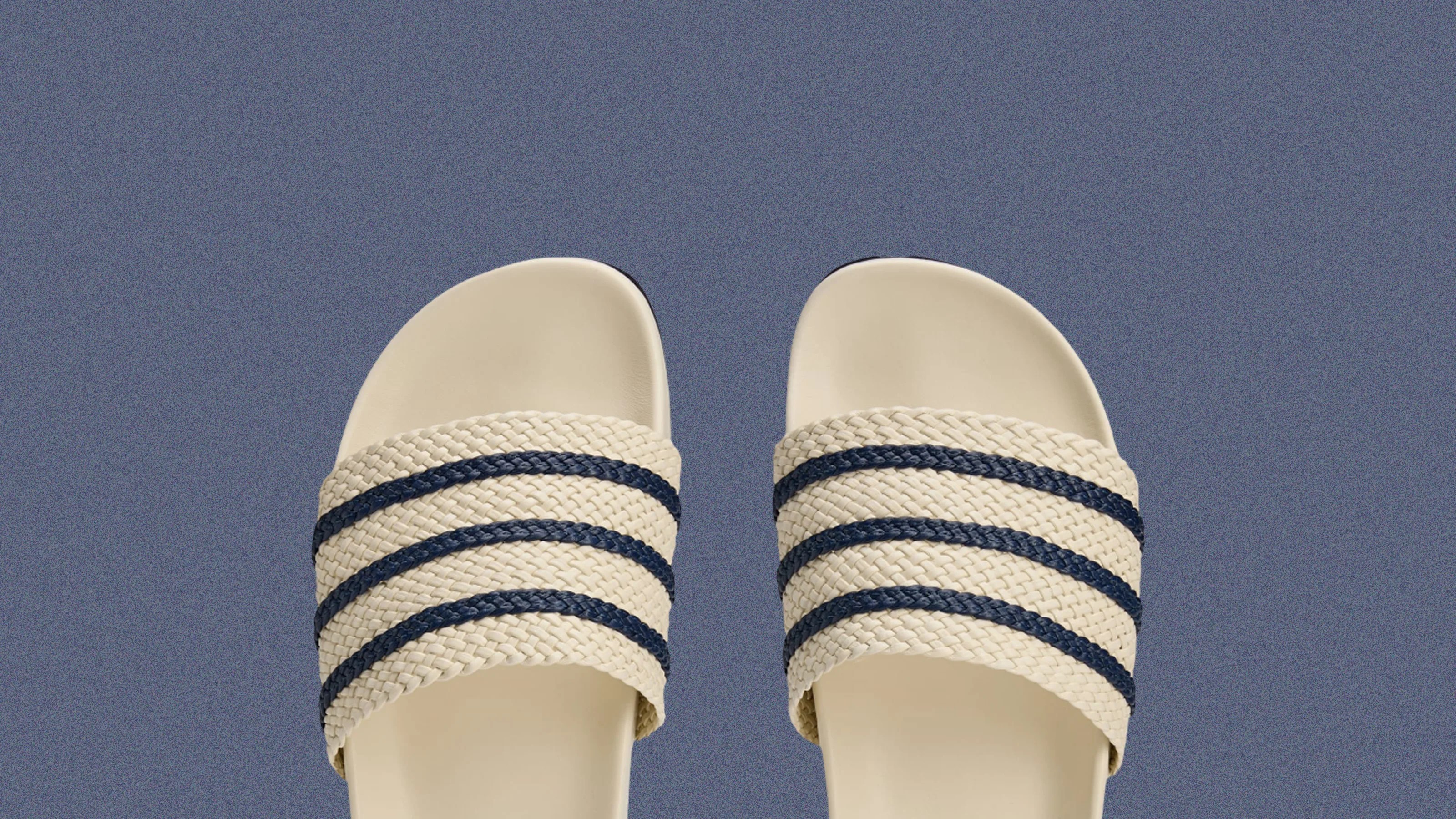 A pair of Wale Bonner Adidas Adilette recovery sandals in cream white with blue strips shown peeking out from the bottom edge of an image with a speckled blue background