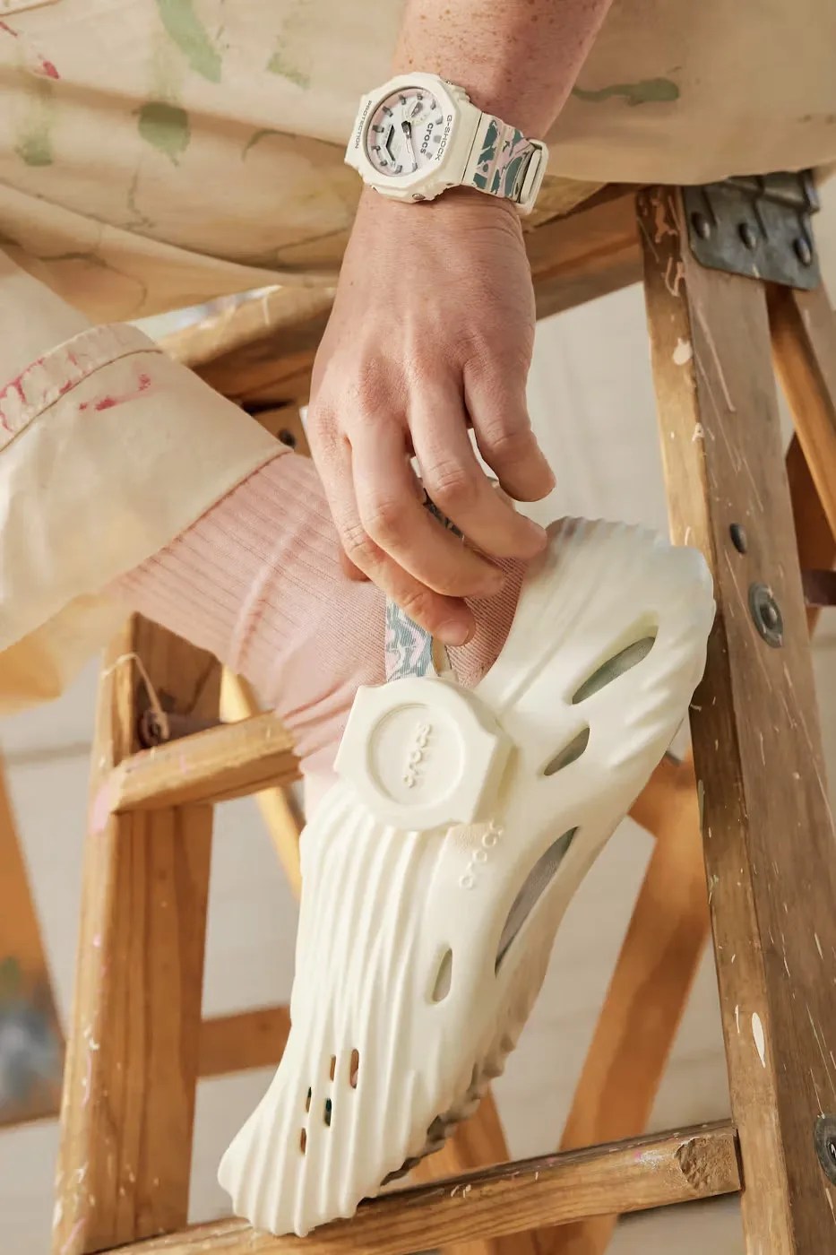 A man is shown wearing stained khaki pants sitting on a stool from the wrist down, wearing the watch and sneakers included in the G-Shock x Crocs Echo Wave watch