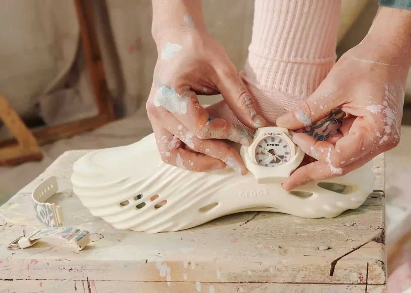 A view of the watch unit of the G-Shock x Crocs Echo Wave watch being slid into the attachment case located on the Crocs Echo Wave shoe by a pair of messy hands covered with specks of white paint. 