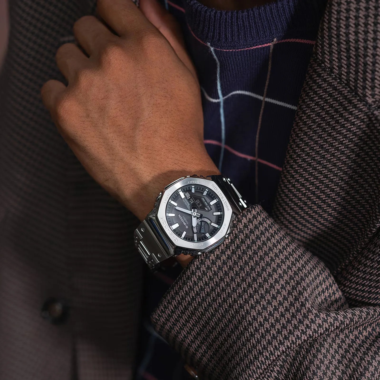 An all-metal version of the G-Shock GA2100 is being worn on a male model's wrist. The model is wearing a herringbone patterned blazer and check patterned navy sweater. 