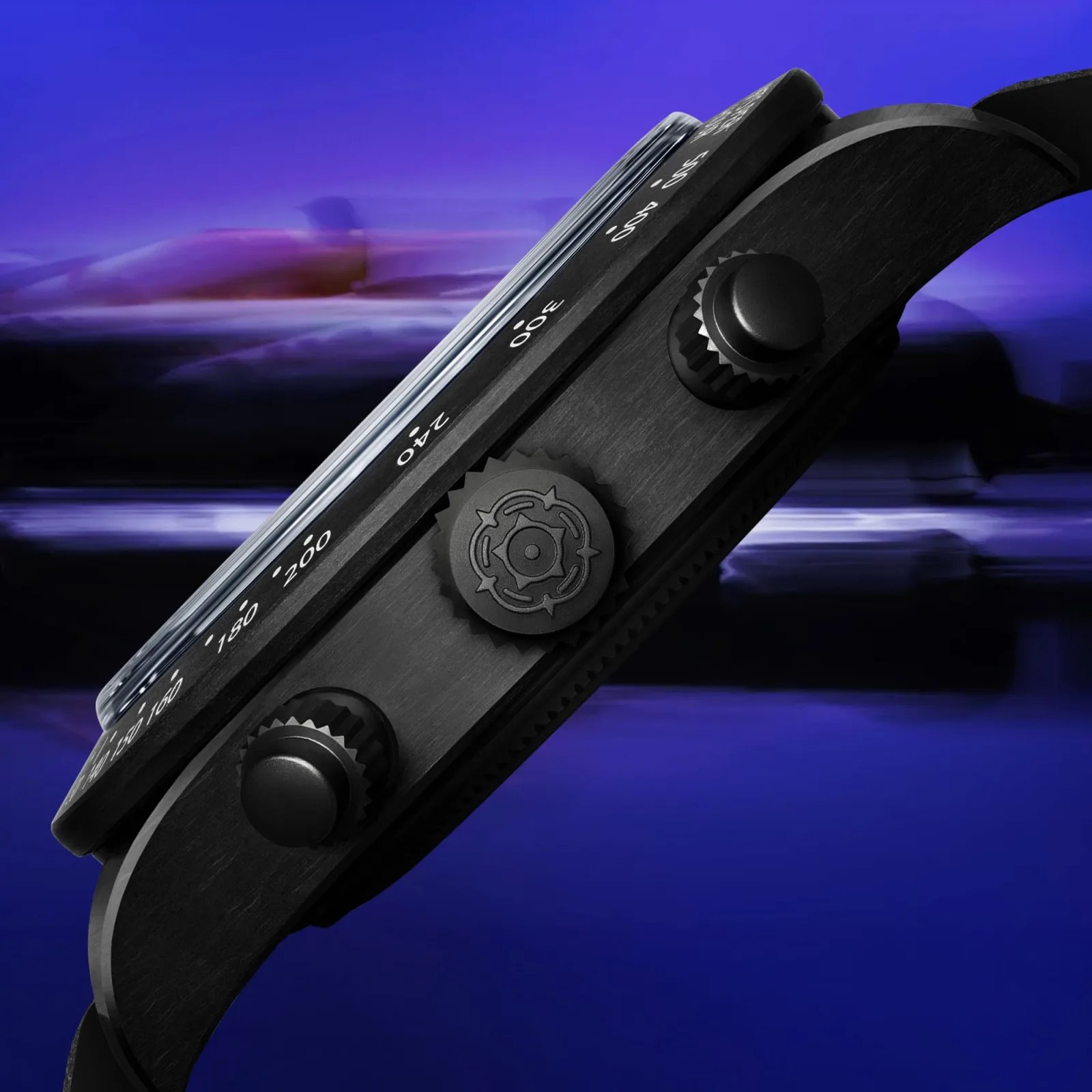 A side few of the crown and pushers of the Tudor Black Bay Chrono "Carbon 25" watch with a blurred F1 car in the background