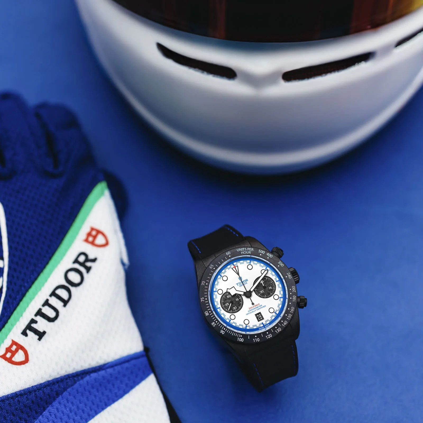 The Tudor Black Bay Chrono "Carbon 25" watch is shown lying on a blue-colored table next to a Tudor-branded racing glove and F1 helmet.