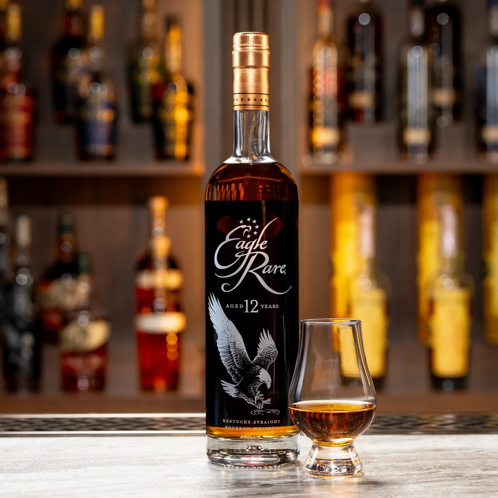 A bottle of Eagle Rare 12 next to a glass of bourbon sitting on a bar backed by other bourbon bottles in the background.