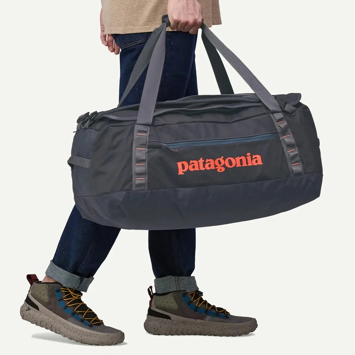Black and dark gray Patagonia duffel bag with orange logo held by a person wearing rolled-up dark jeans and hiking shoes.