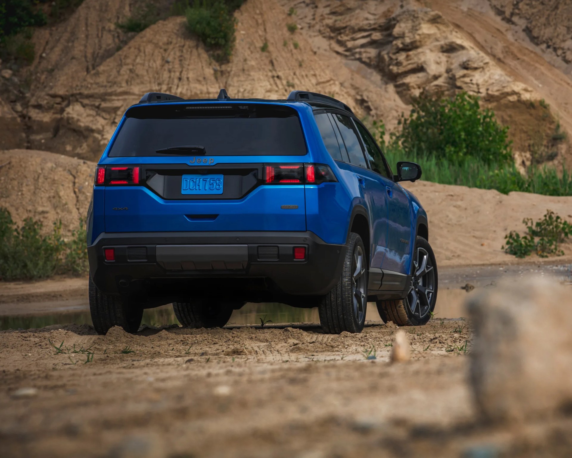 Blue Jeep SUV parked on a sandy, rocky terrain with a natural backdrop of dirt hills and greenery, viewed from the rear. The vehicle has tinted rear windows, black lower bumper trim, and modern taillights. The license plate reads "DCH 758.