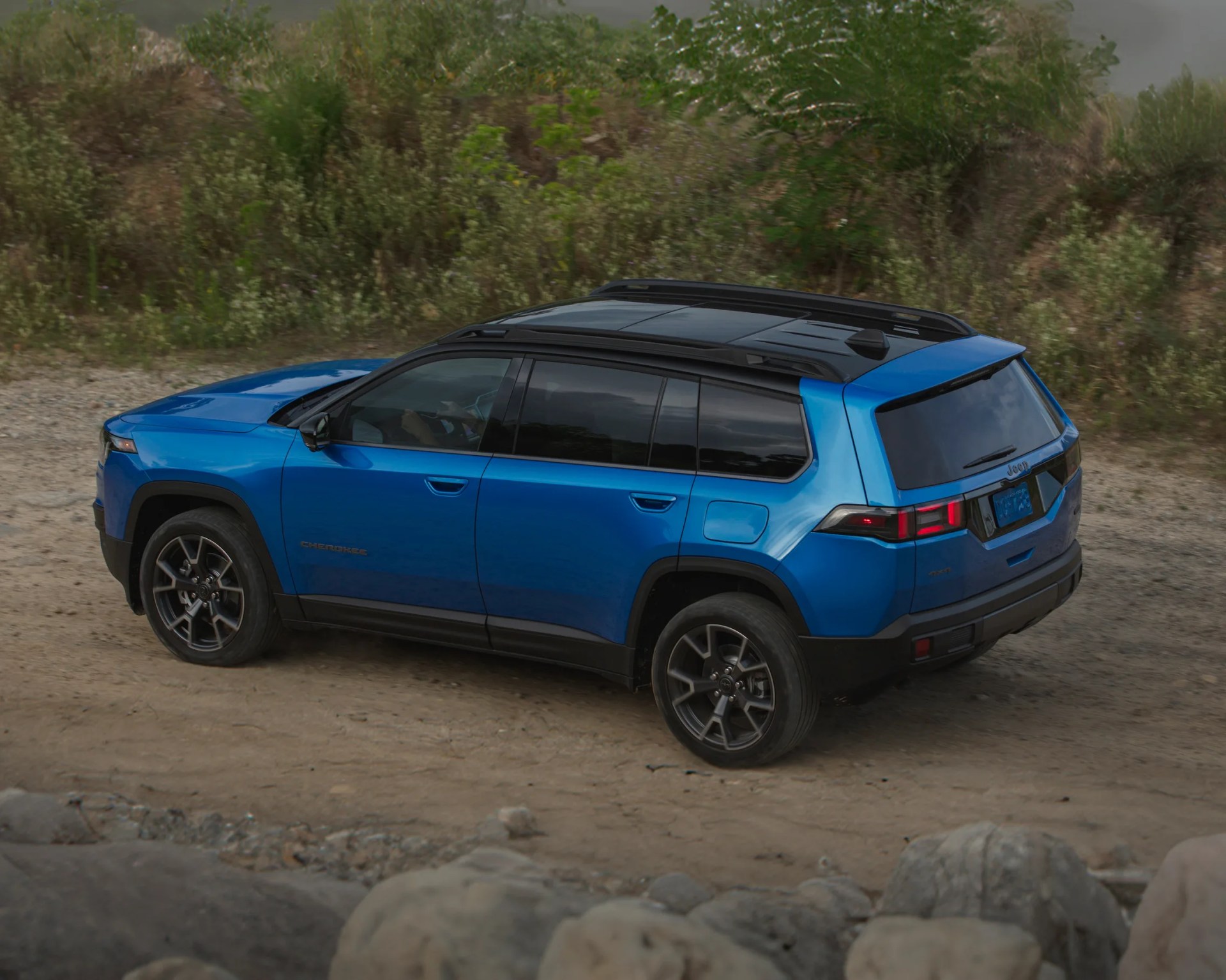 Blue Jeep Cherokee SUV with black roof rails and tinted windows parked on a dirt path, surrounded by greenery and rocks.