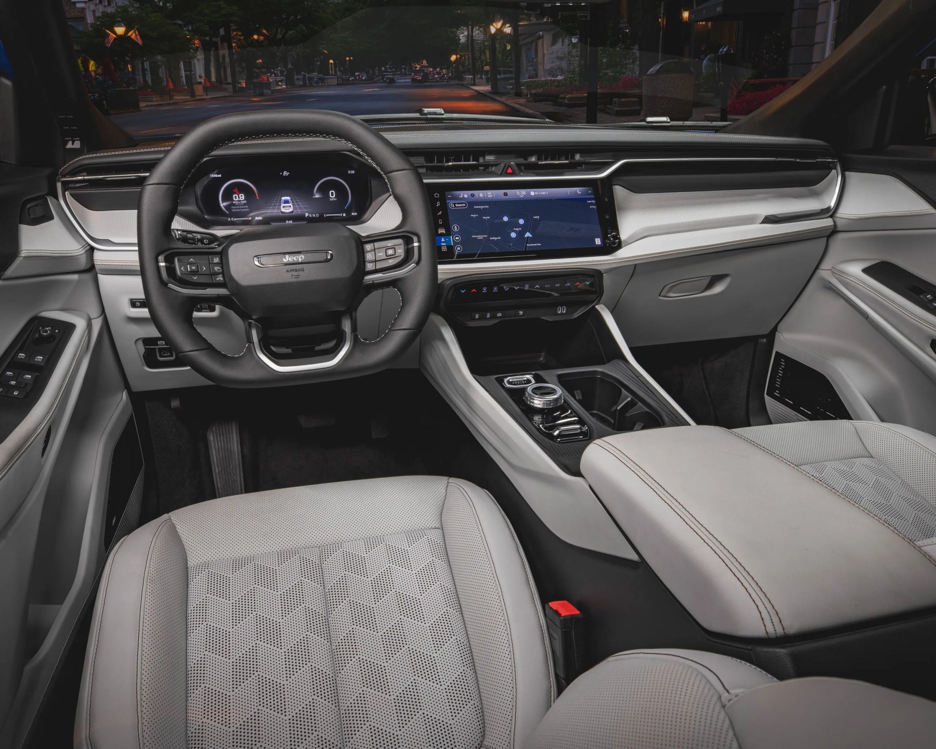 Interior of a Jeep vehicle featuring a black leather-wrapped steering wheel with control buttons, a digital instrument cluster, and a large touchscreen infotainment system displaying a navigation map. The cabin has light gray perforated leather seats, a center console with a rotary gear selector, cup holders, and climate control buttons below the screen. The dashboard and door panels combine light gray and black materials with contrast stitching.