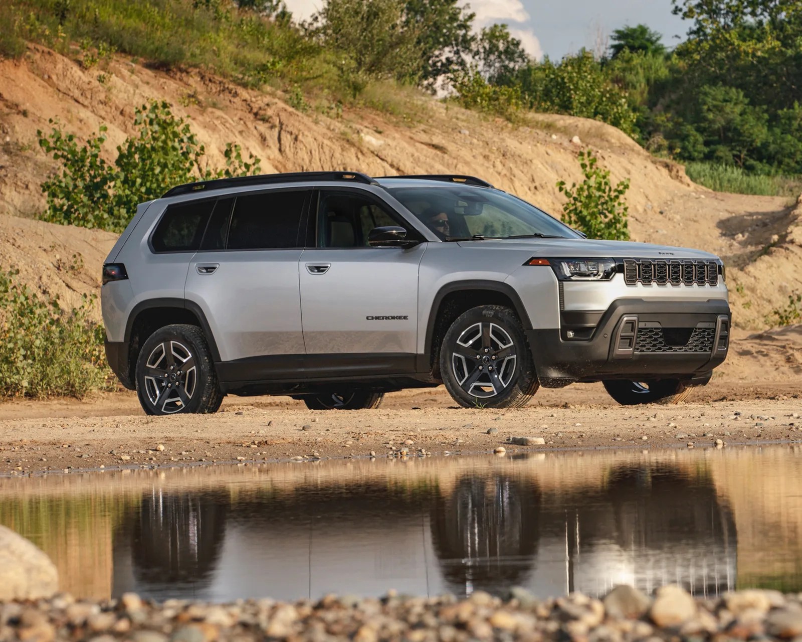Silver Jeep Cherokee SUV parked on a sandy area near a small body of water, with greenery and rocky terrain in the background. The vehicle has black trim around the wheel arches and lower body, modern alloy wheels, and a distinctive front grille. The reflection of the SUV is visible in the water.