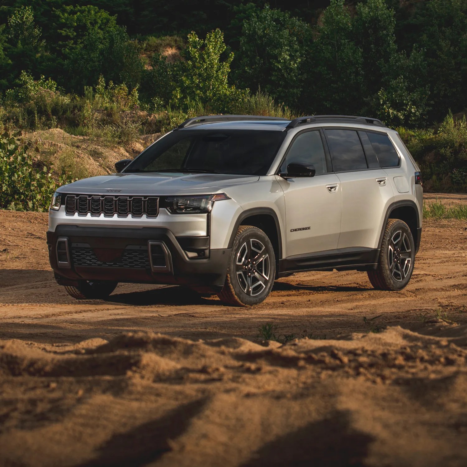 Silver Jeep Cherokee SUV parked on a dirt surface with green trees in the background, featuring black trim, tinted windows, and modern alloy wheels.