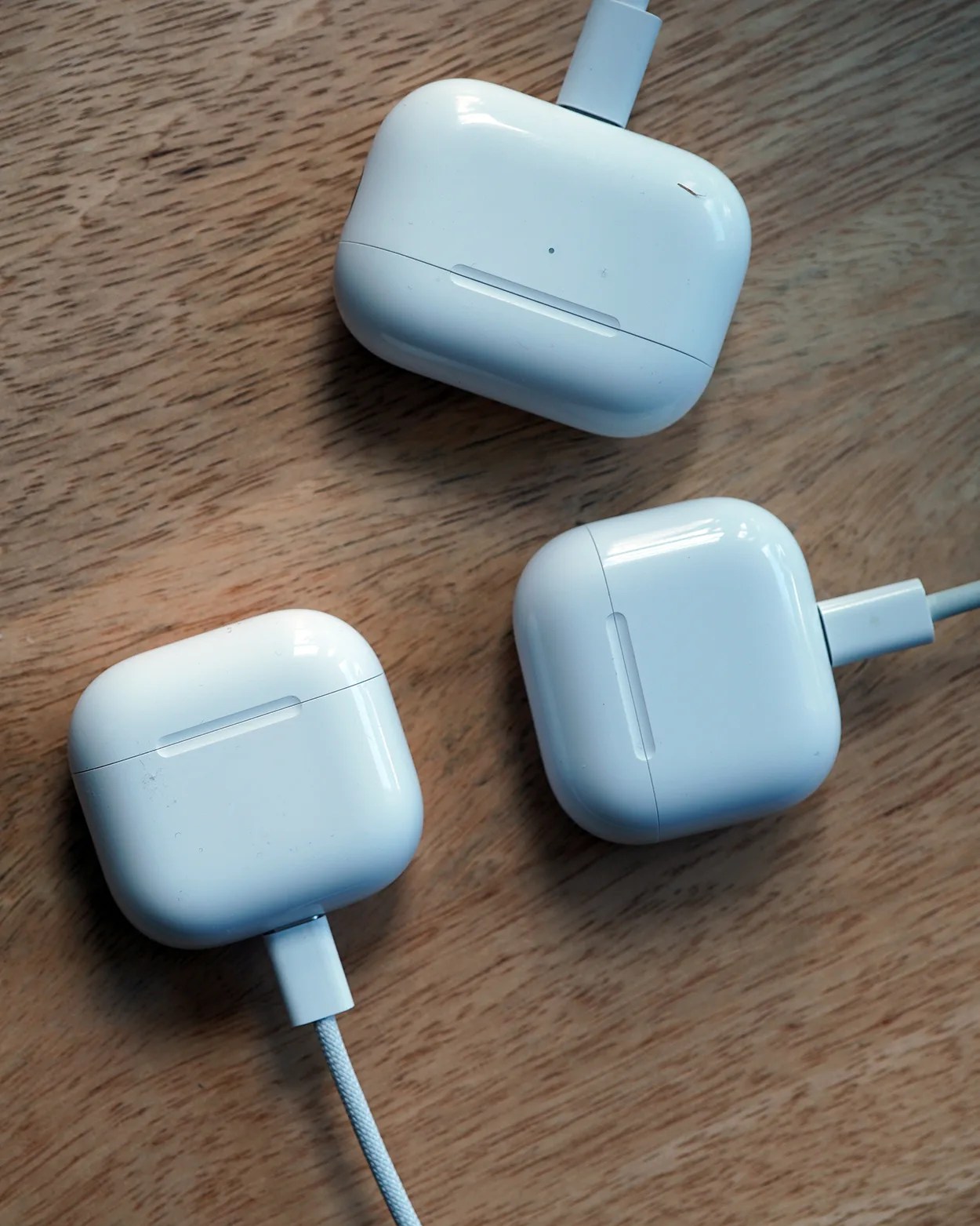 Three white wireless earbud charging cases are shown on a wooden surface, each connected to a charging cable. The cases have a glossy finish and rounded edges.