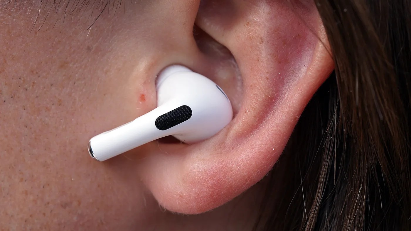 Close-up of a person's ear with a white wireless earbud inserted, featuring a black mesh area on the stem. The skin around the ear shows natural texture and some small blemishes. Dark hair is visible on the right side.