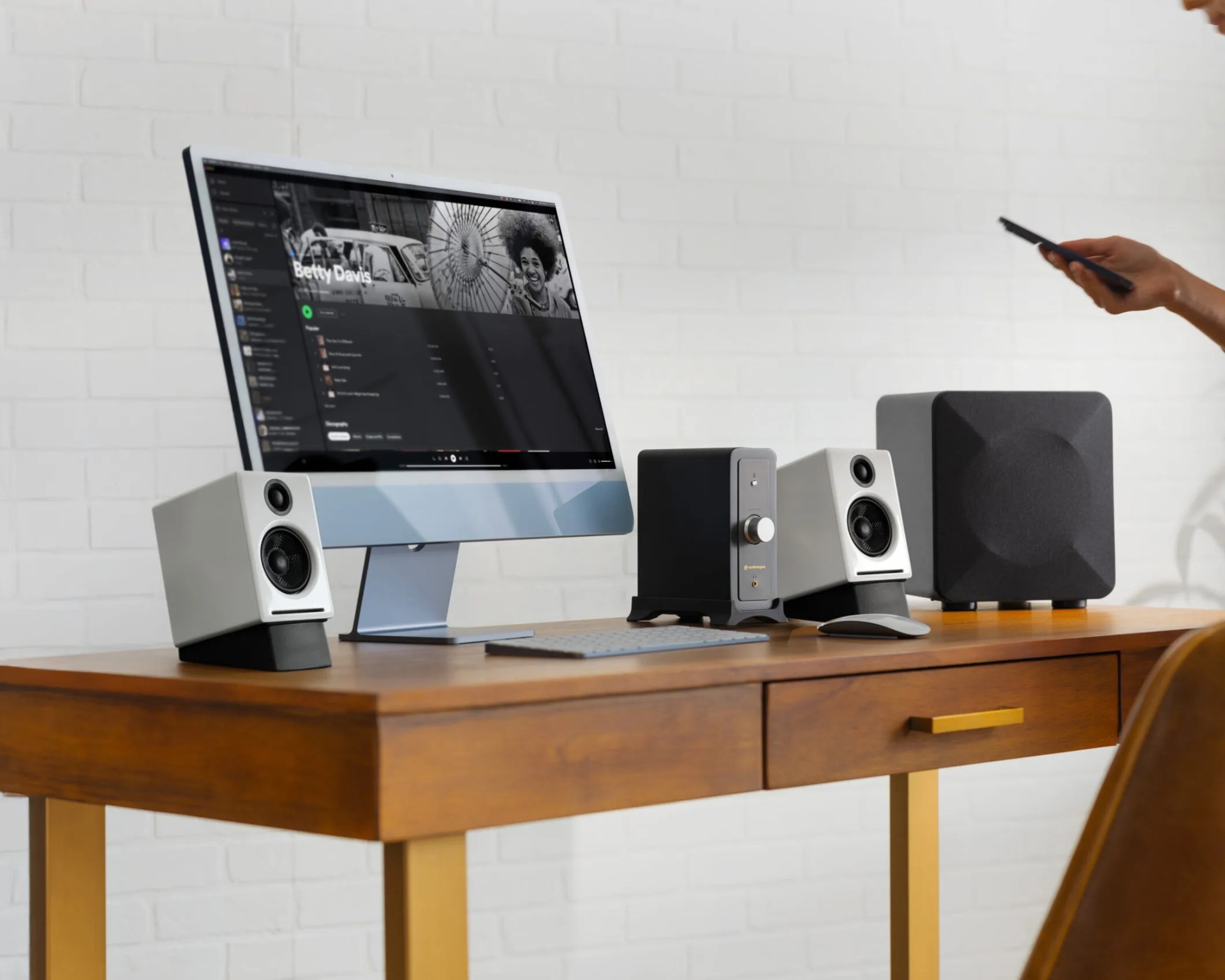 A wooden desk with gold-colored legs holds a modern all-in-one computer displaying a music playlist. On the desk are two white speakers with black stands, a black audio amplifier with a silver knob, a wireless keyboard, a wireless mouse, and a large black subwoofer. A person is holding a smartphone on the right side of the image. The background features a white brick wall.